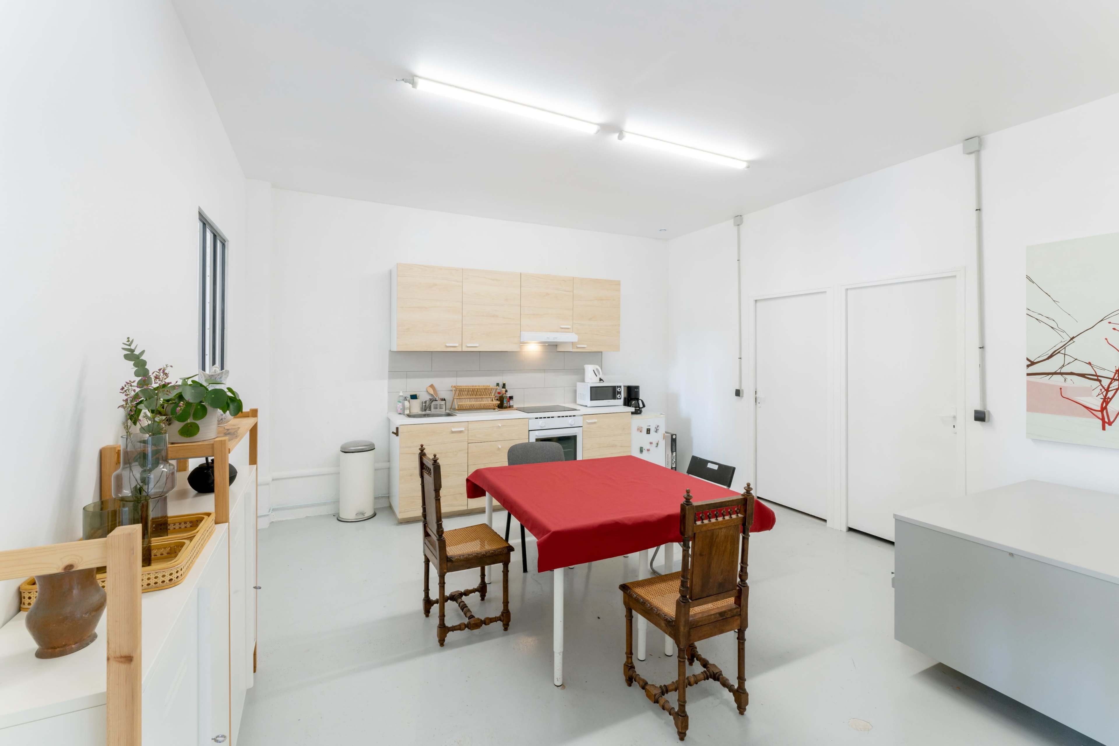 A simple kitchen with light wood cabinets, a red table in the center, and four chairs, featuring a microwave and white appliances.