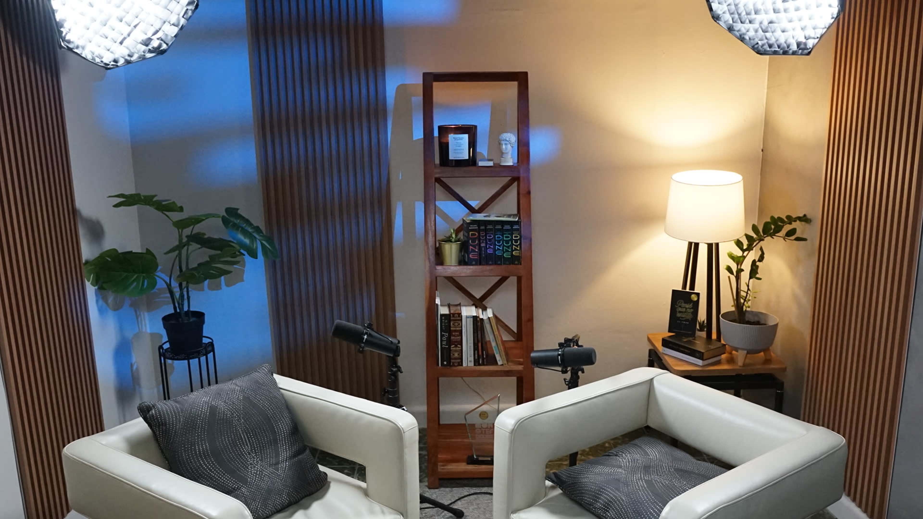 The image shows a cozy interview setup with two white chairs facing a wooden shelf filled with books, a potted plant, and a lamp in a softly lit room.