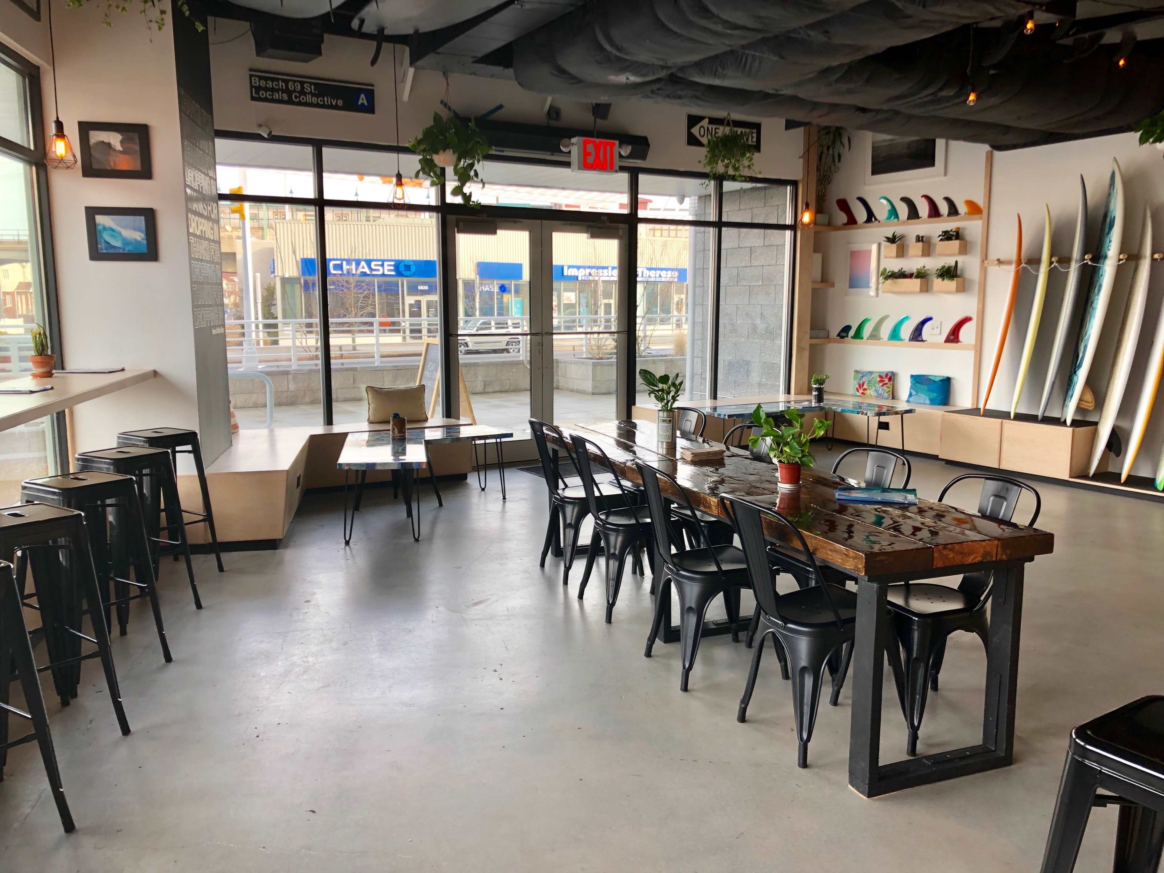 A spacious indoor area features a long wooden table surrounded by black chairs, large windows allowing natural light, and surfboards displayed on the wall.