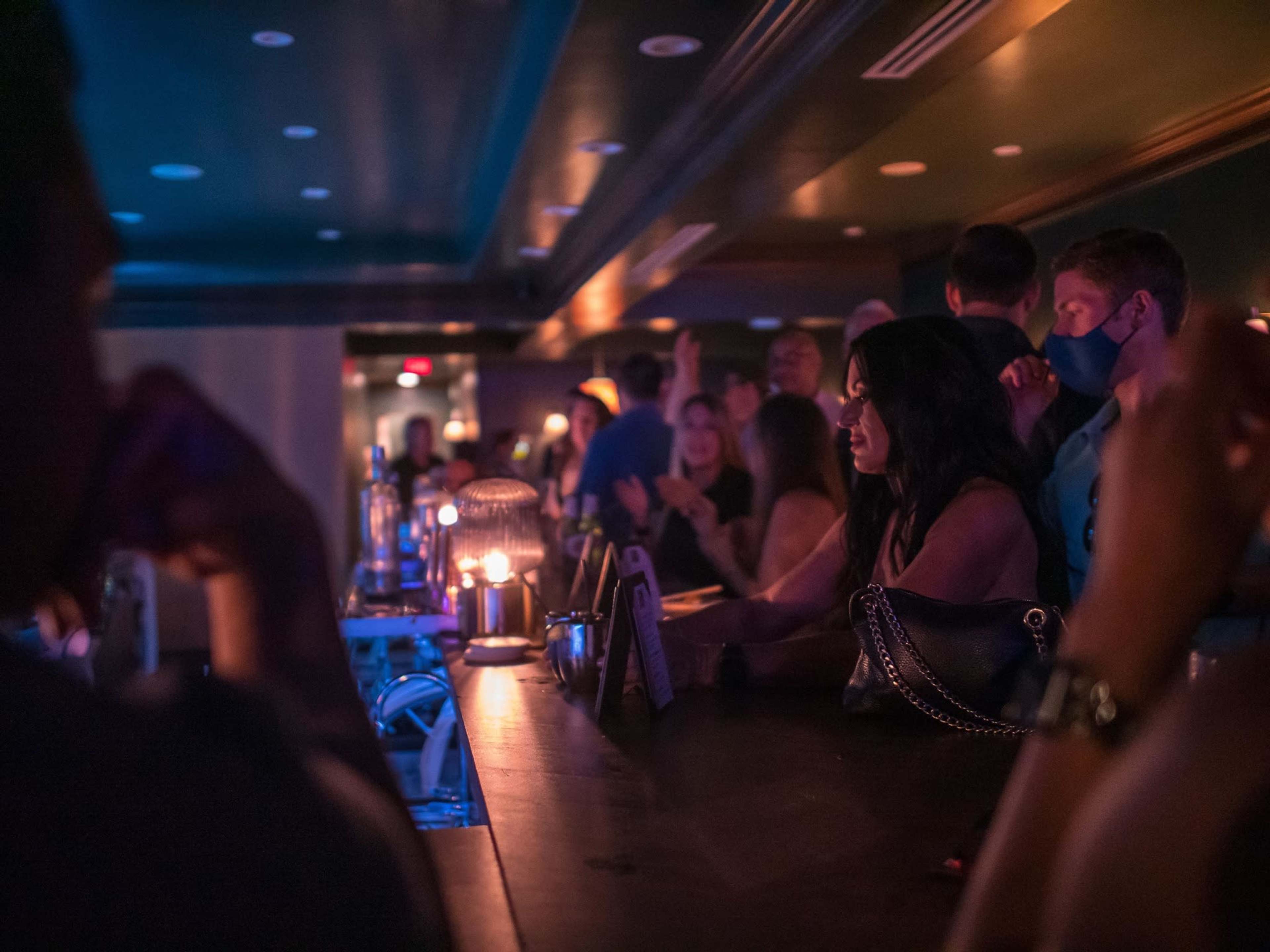 A dimly lit bar scene features patrons socializing, with one woman resting her chin on her hand while wearing a mask.
