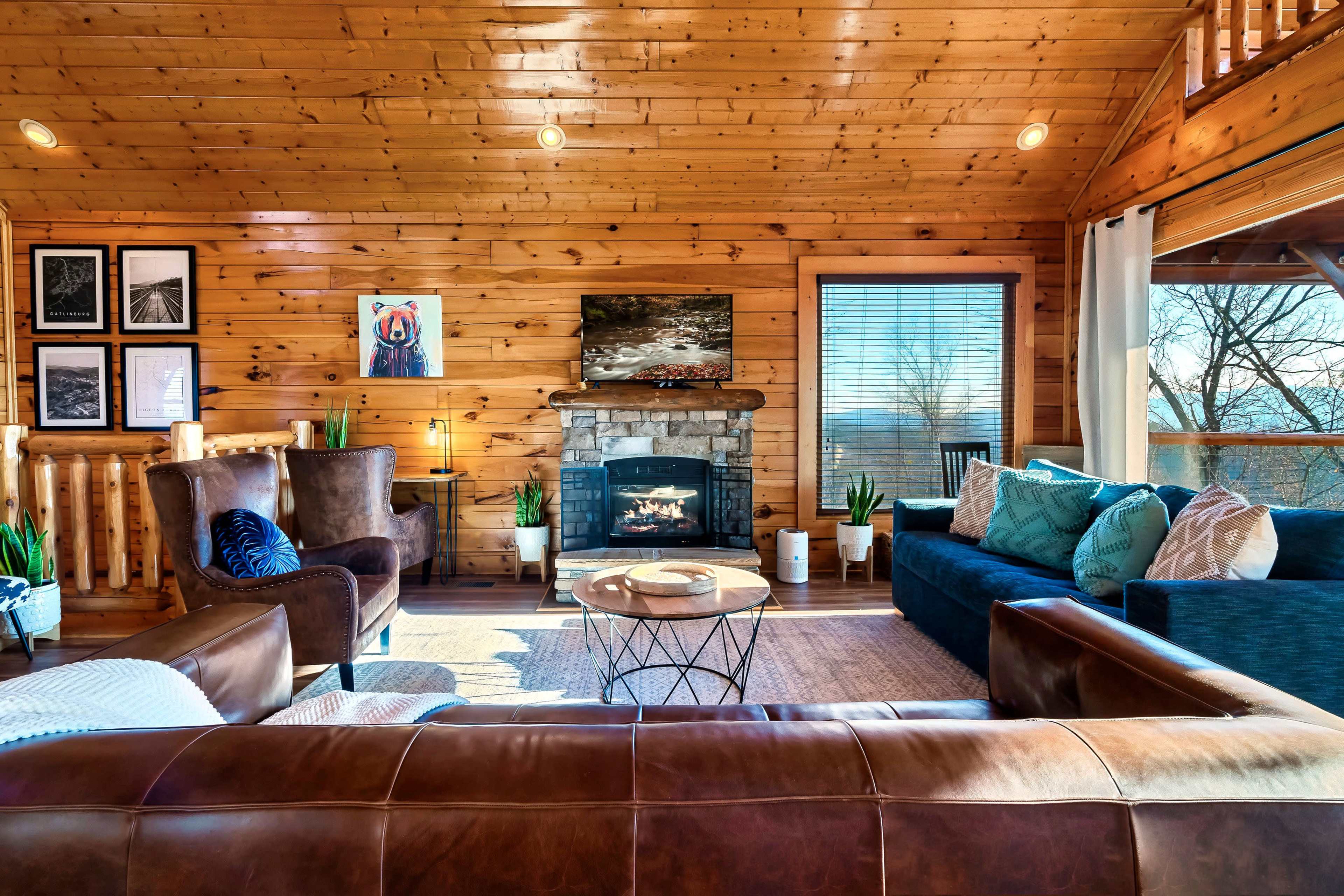 The image shows a warm and inviting wooden cabin interior with a stone fireplace, comfortable seating, and large windows revealing a scenic view.