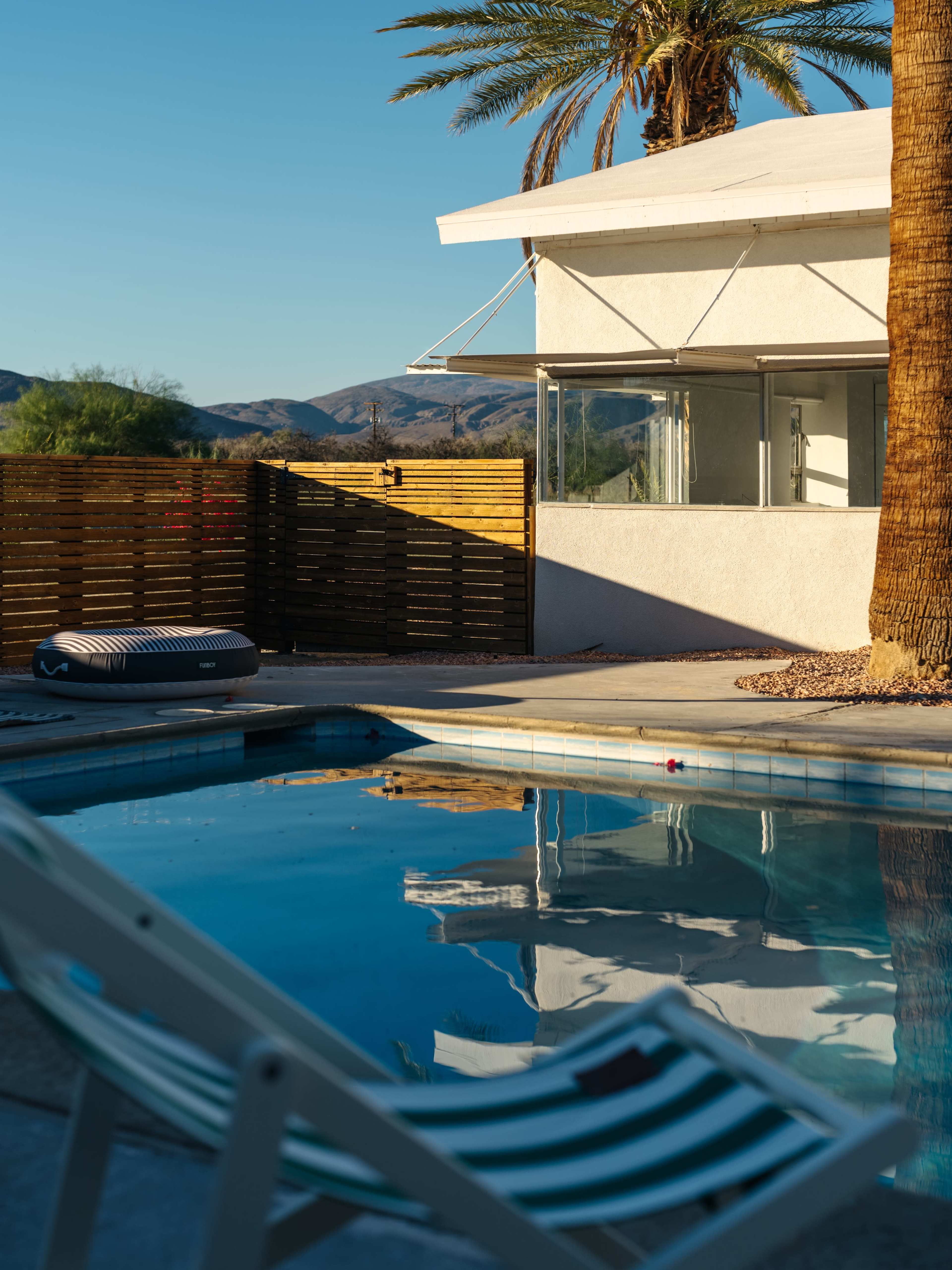 The image shows a swimming pool reflecting the surrounding palm trees and a modern building, with a lounge chair positioned nearby.