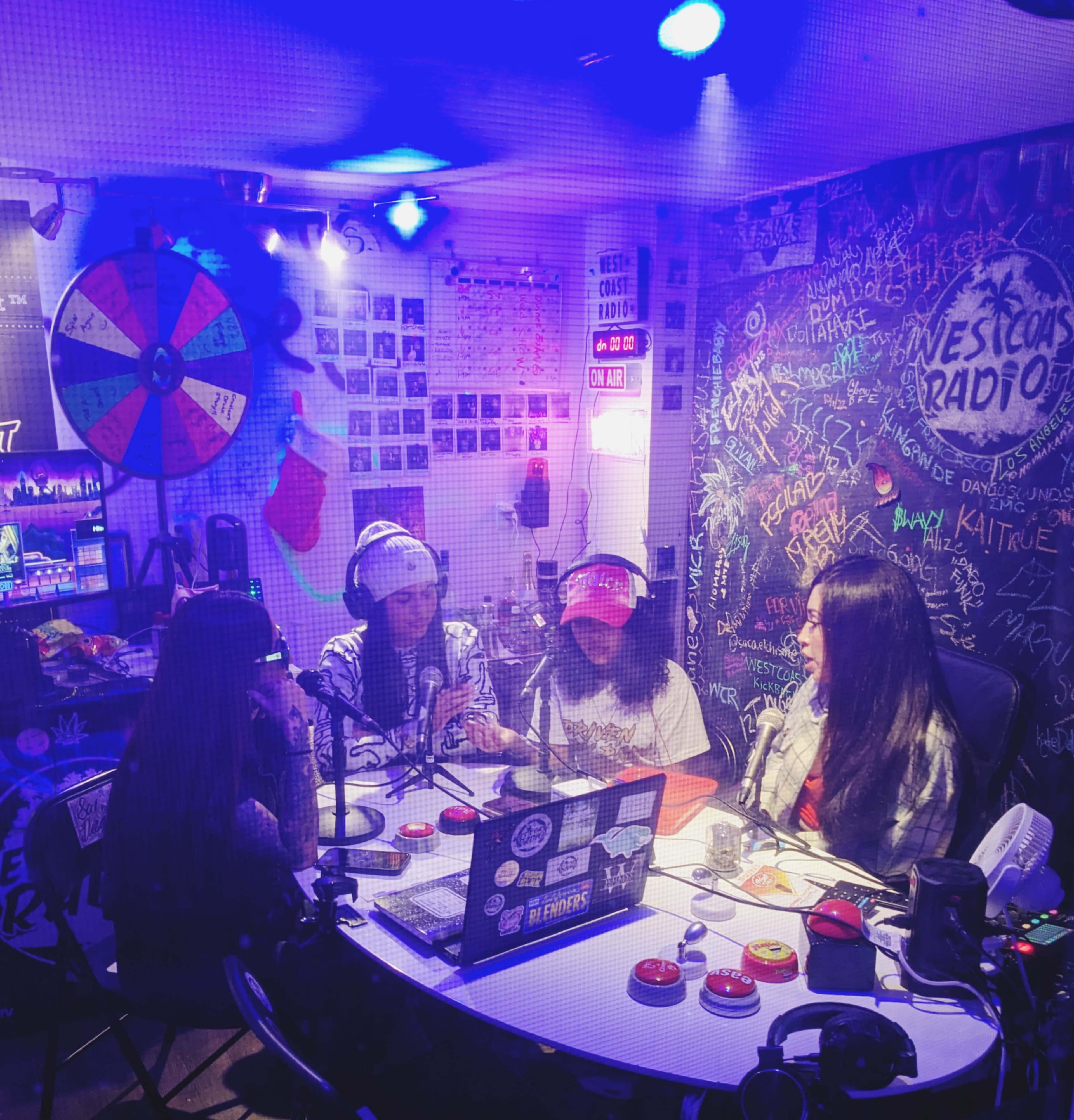 Four individuals are seated around a table in a radio studio, engaged in conversation, with microphones and various colorful decorations surrounding them.