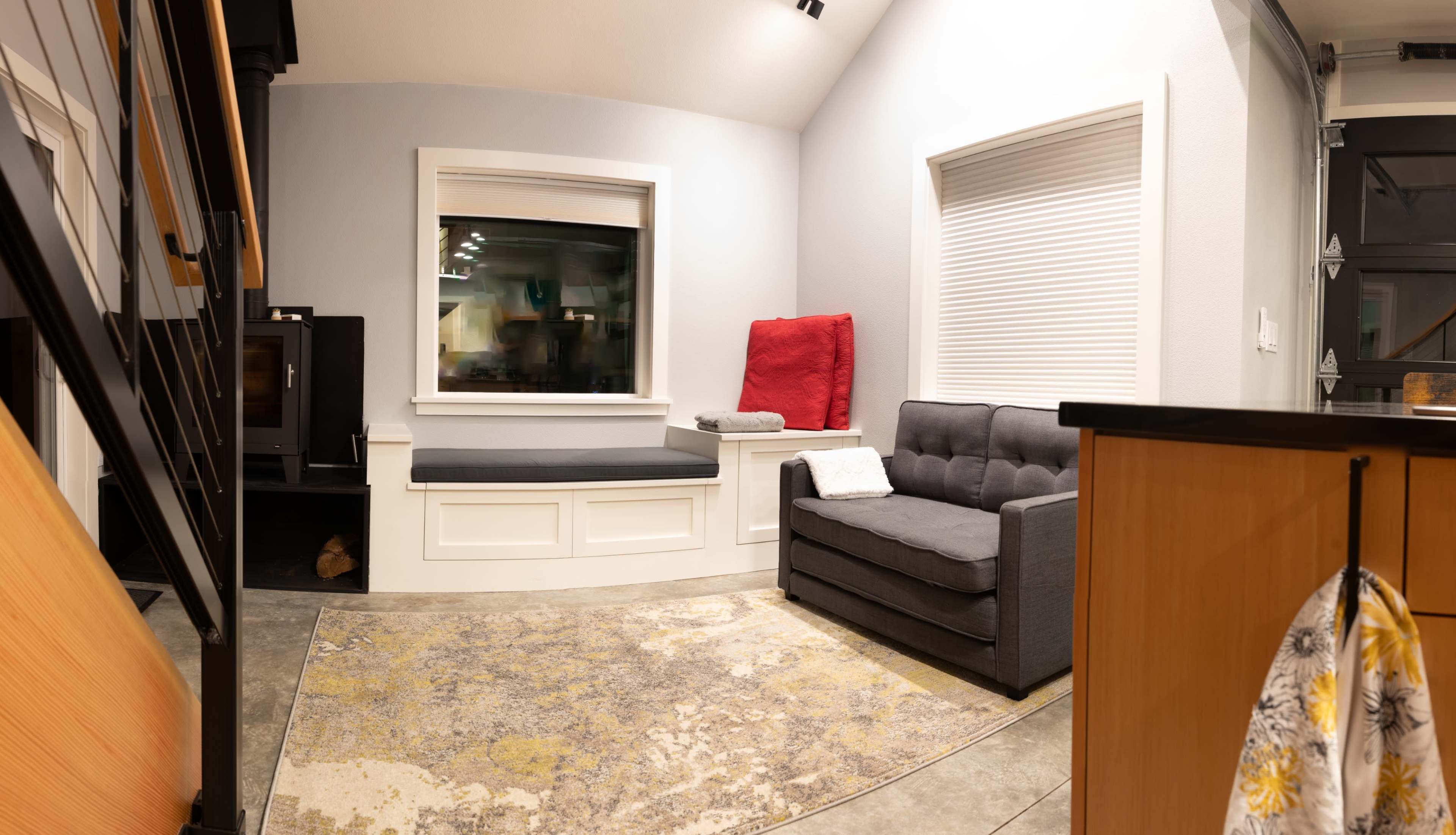 The image shows a cozy interior space featuring a gray sofa, a window seat with cushions, and a rug on a concrete floor.