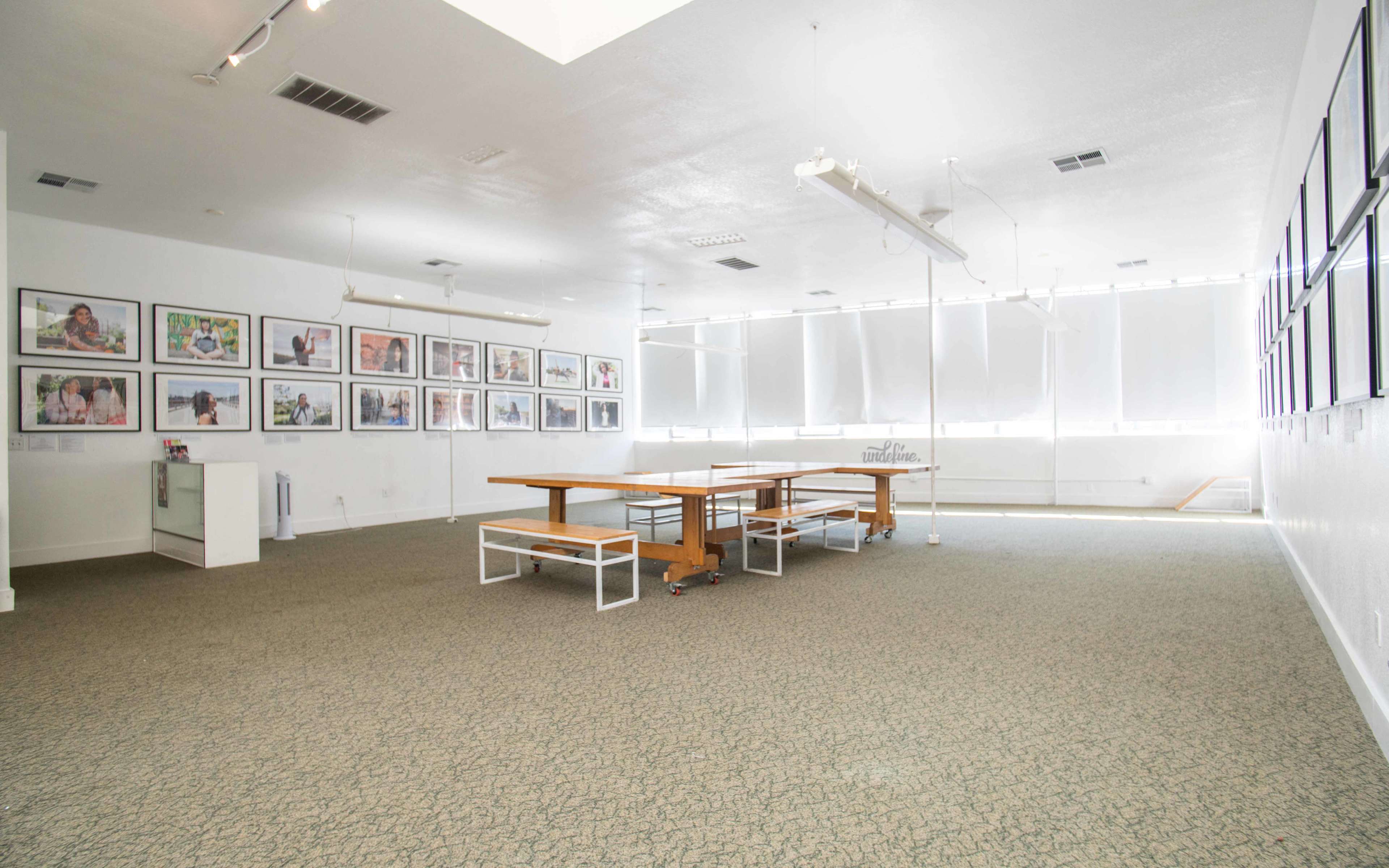A spacious, brightly lit room features a central wooden table surrounded by benches, with framed photographs hung along the white walls.