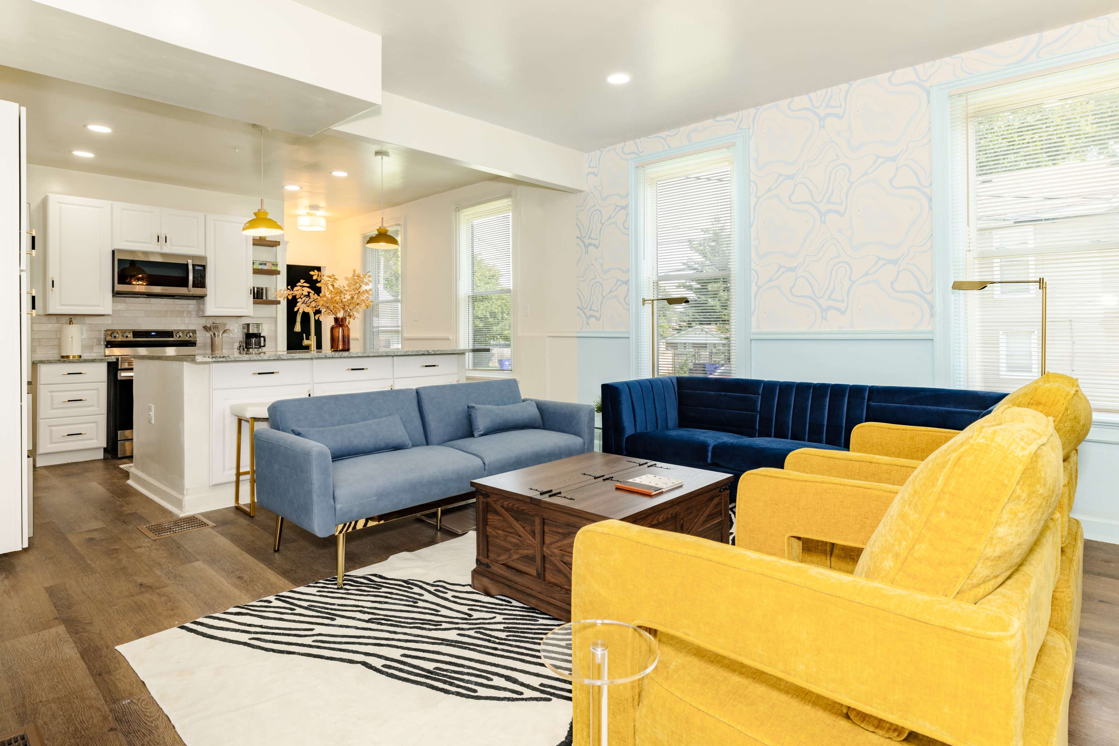 A modern living room features a blue sofa, a yellow sofa, and a wooden coffee table on a patterned rug, with a light-filled kitchen visible in the background.
