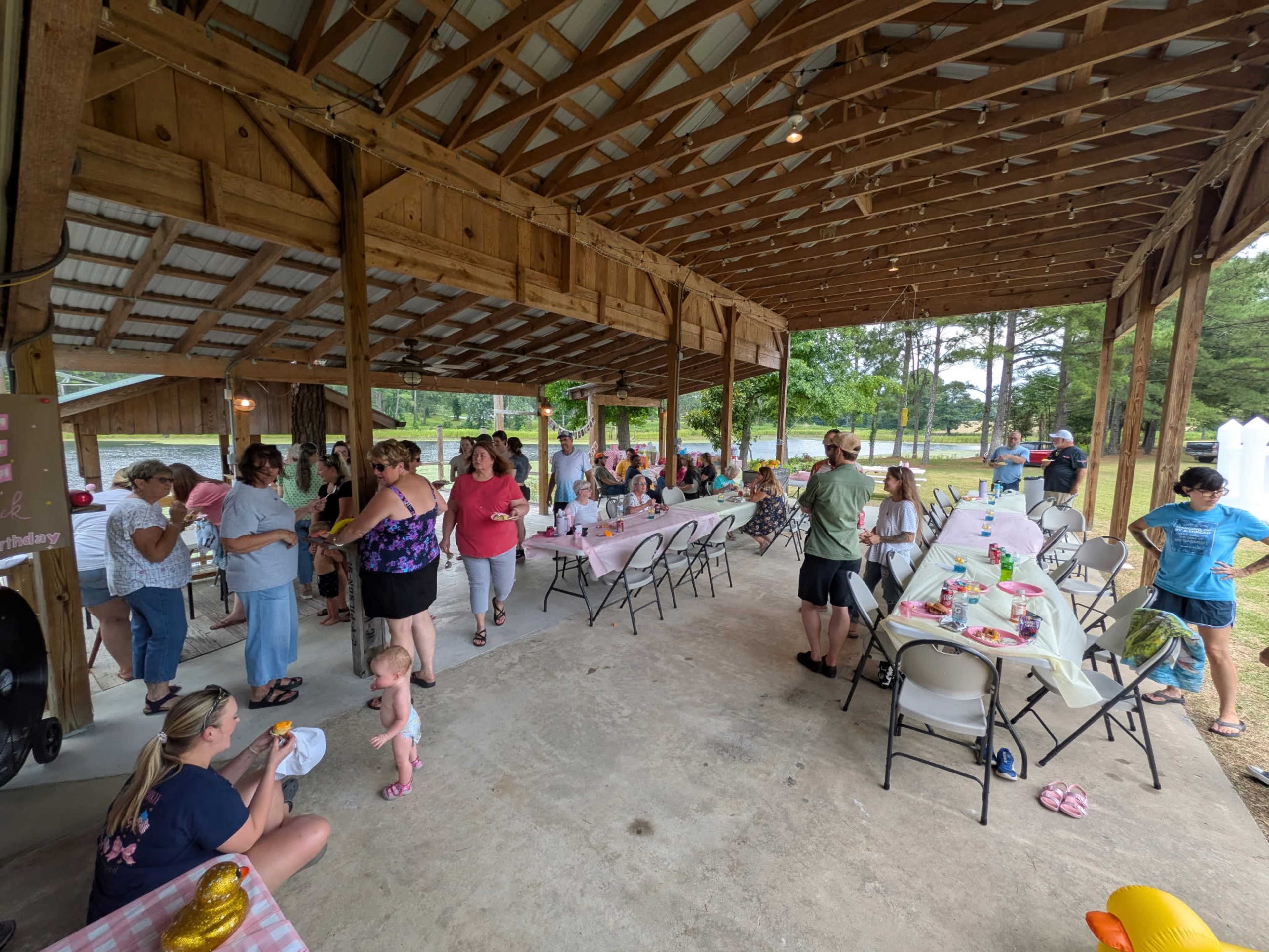 A group of people is gathered under a wooden pavilion for a social event, with tables set up for food and drinks.