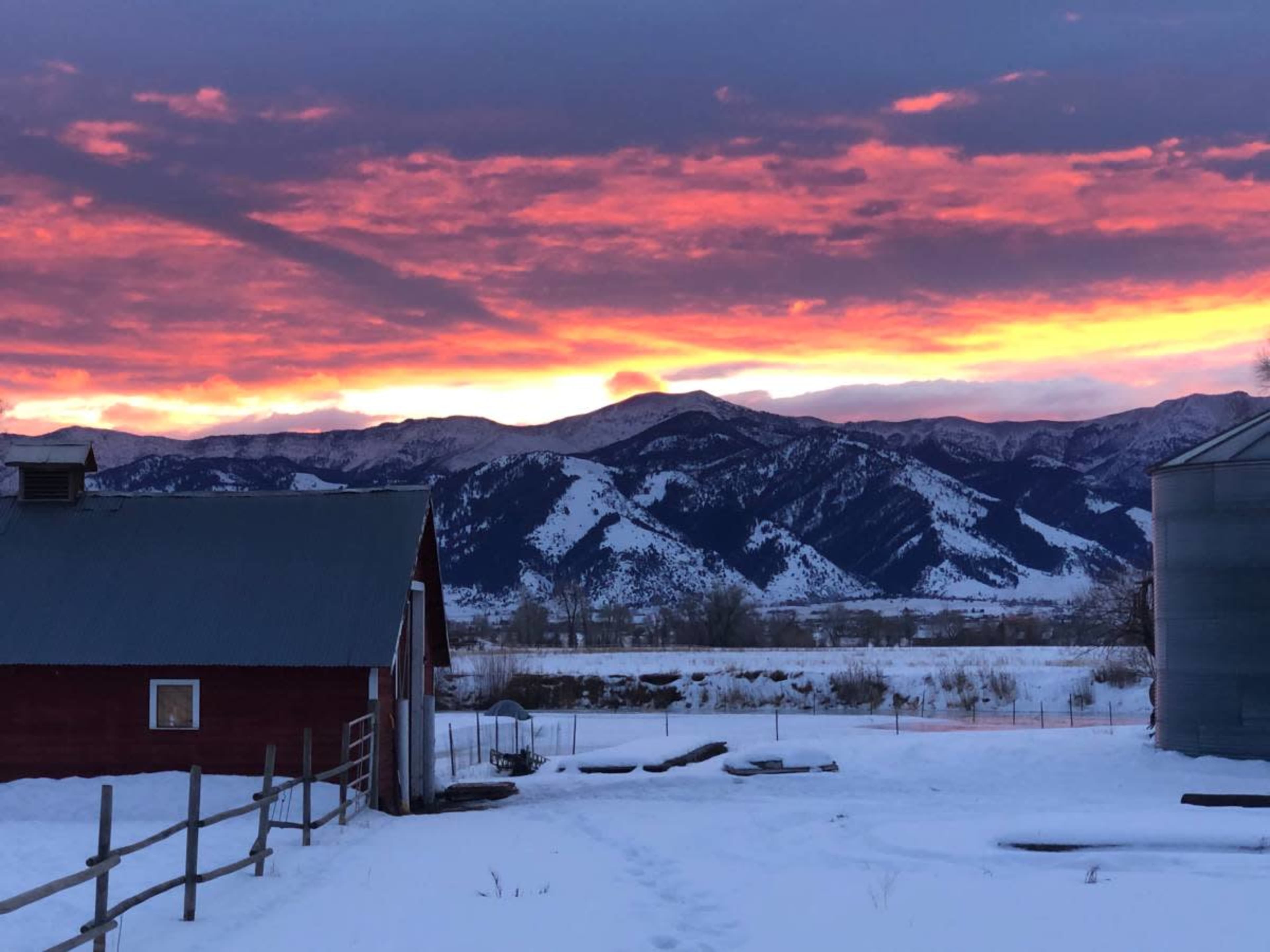 The image shows a red barn and a silos against a backdrop of snow-covered ground and mountains under a colorful sunset.