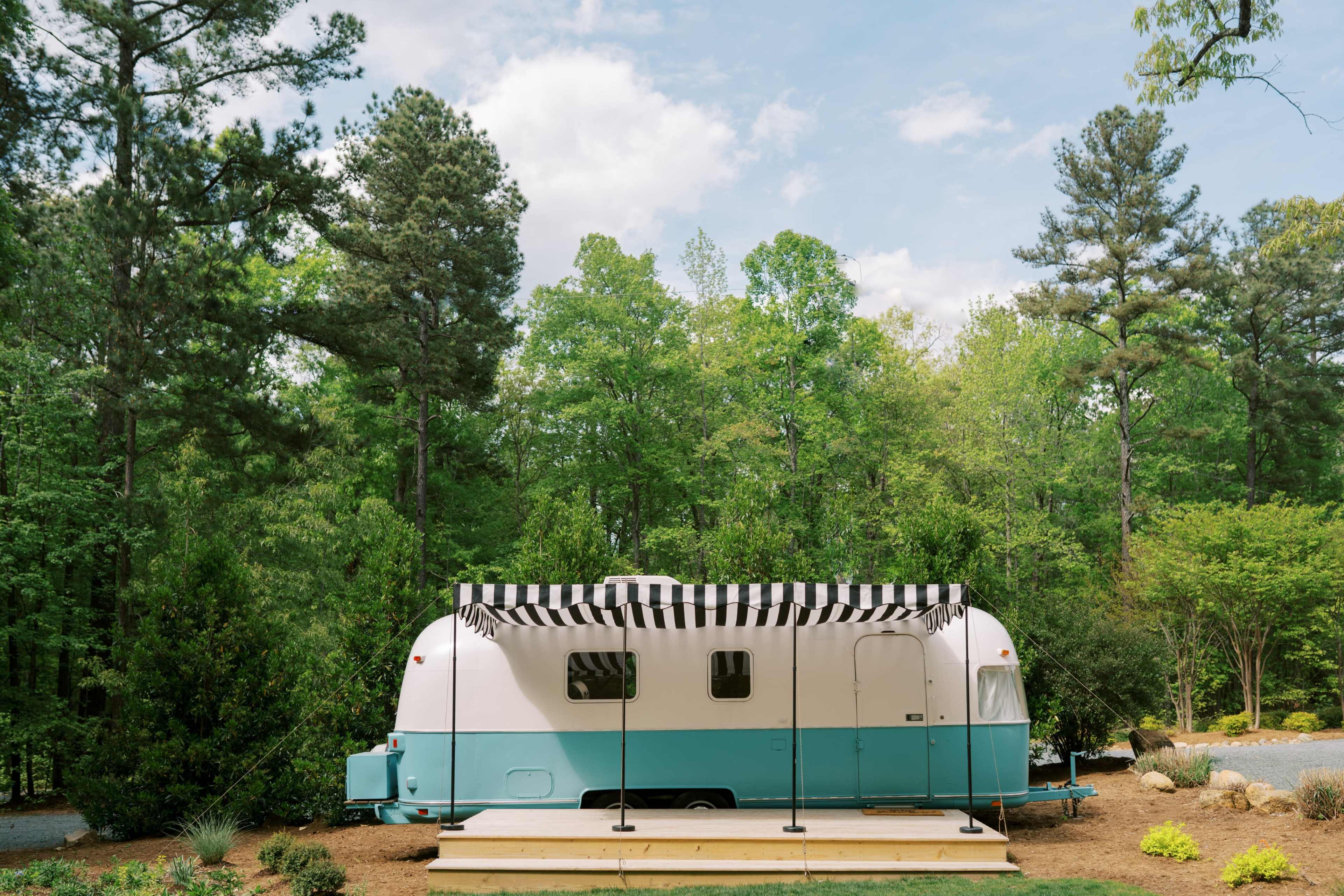 A vintage camper trailer with a blue-and-white exterior is parked in a wooded area, featuring a striped awning above a small wooden deck.