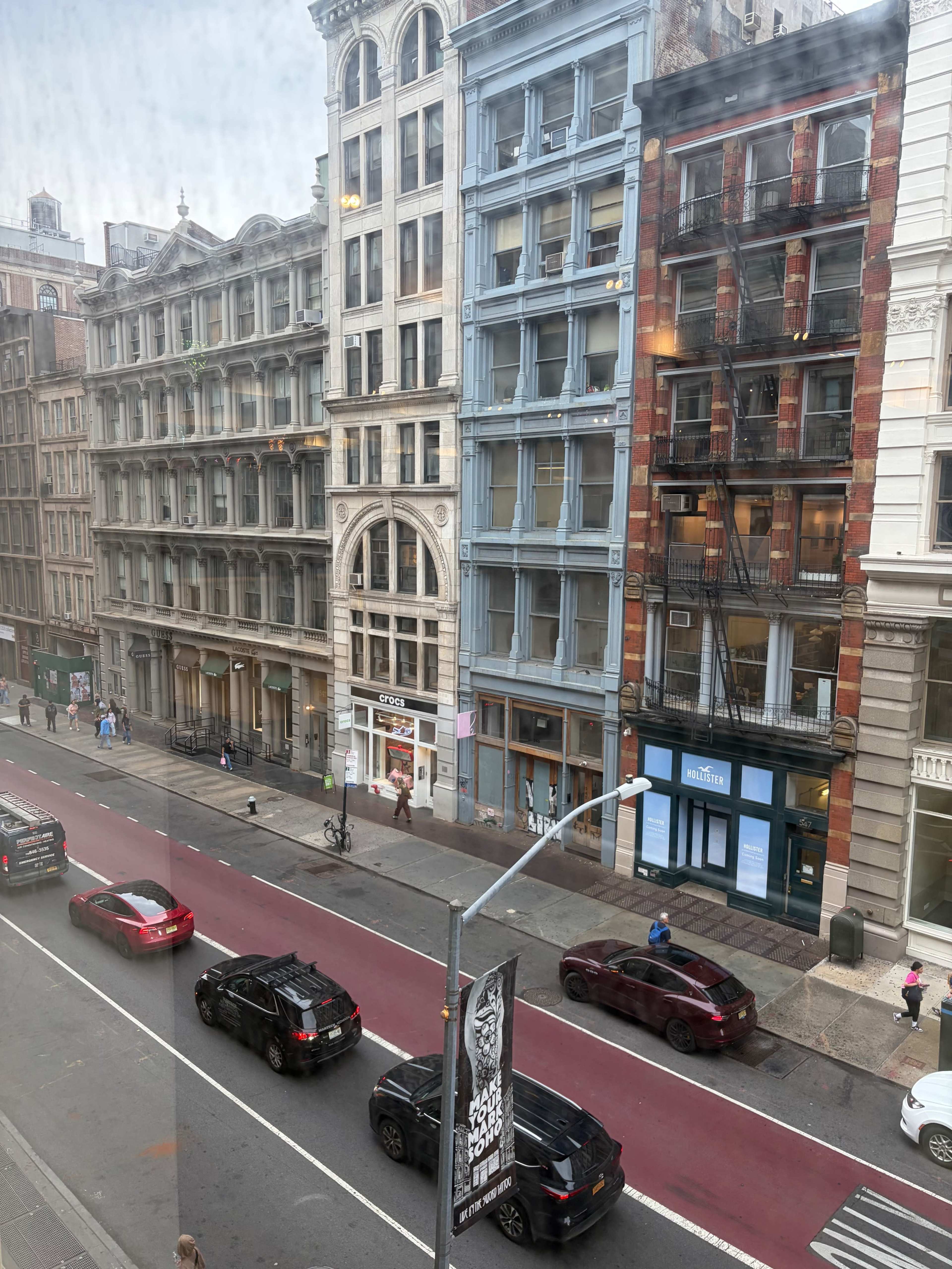 The image shows a street view of buildings with varied architectural styles, featuring multiple stories and storefronts, while cars are parked along a red-lined road.