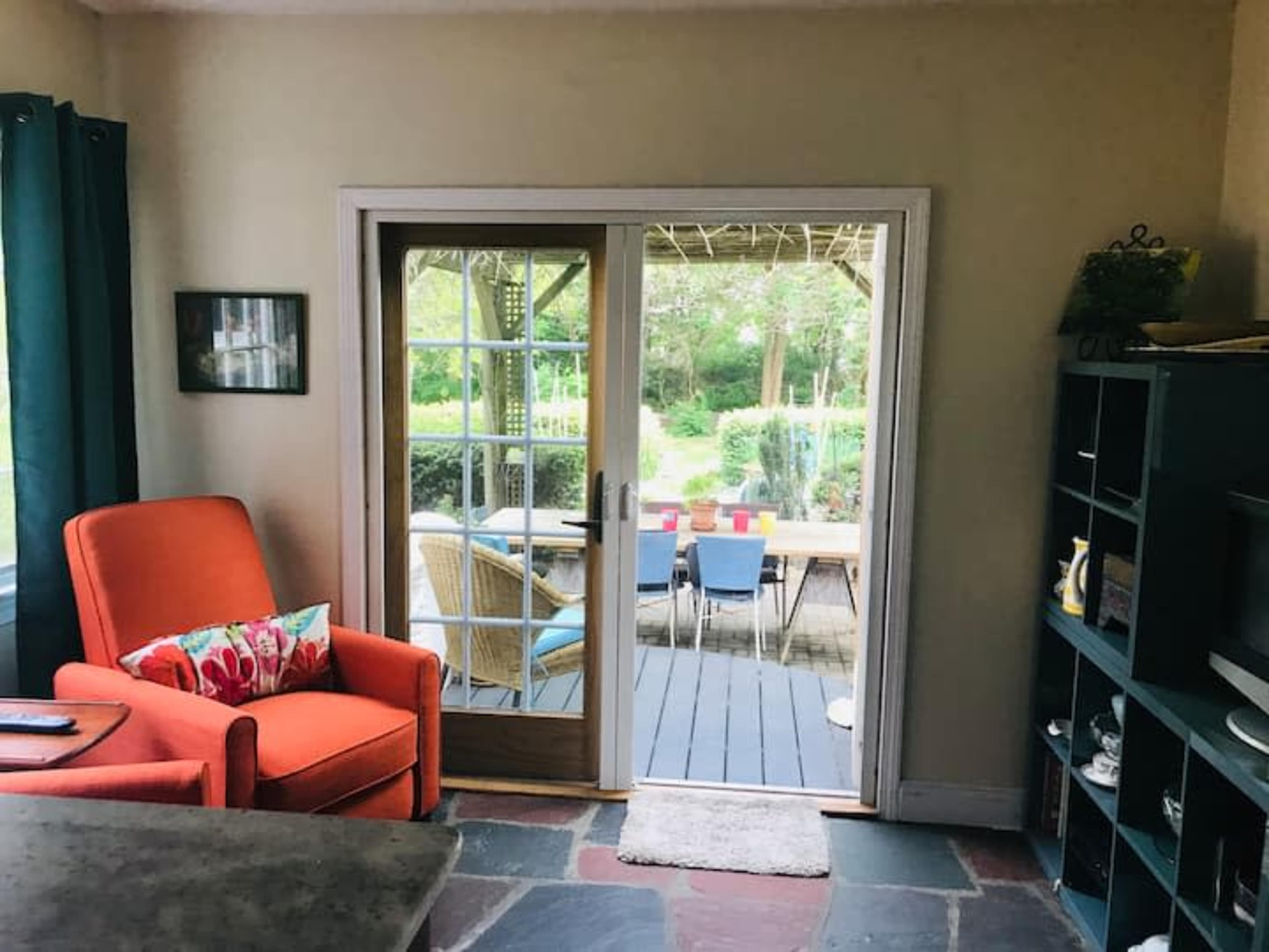 A cozy room features an orange armchair and a sliding glass door that opens to a patio surrounded by greenery.