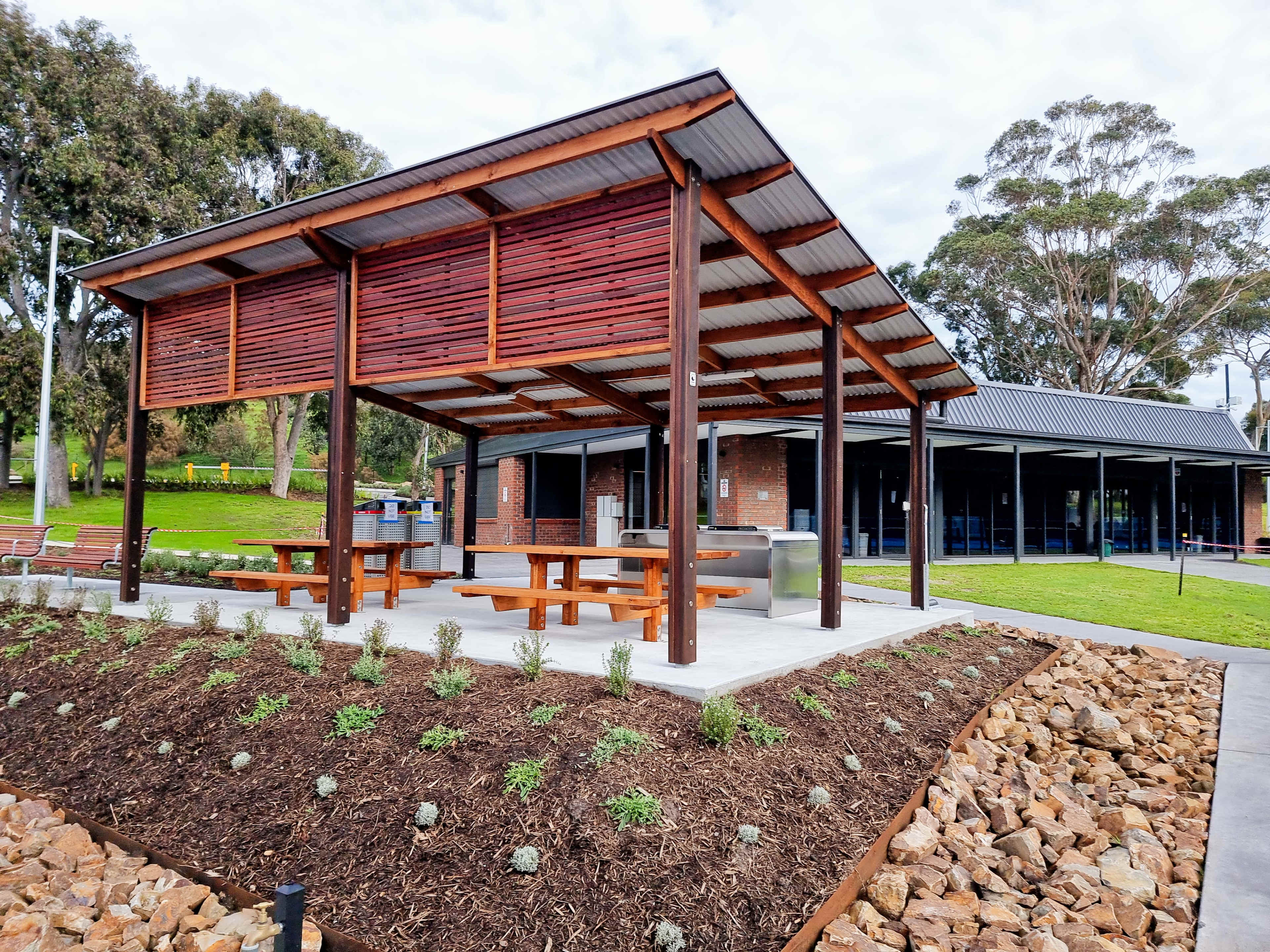 A wooden pavilion with a metal roof, featuring picnic tables and surrounded by landscaped garden beds.