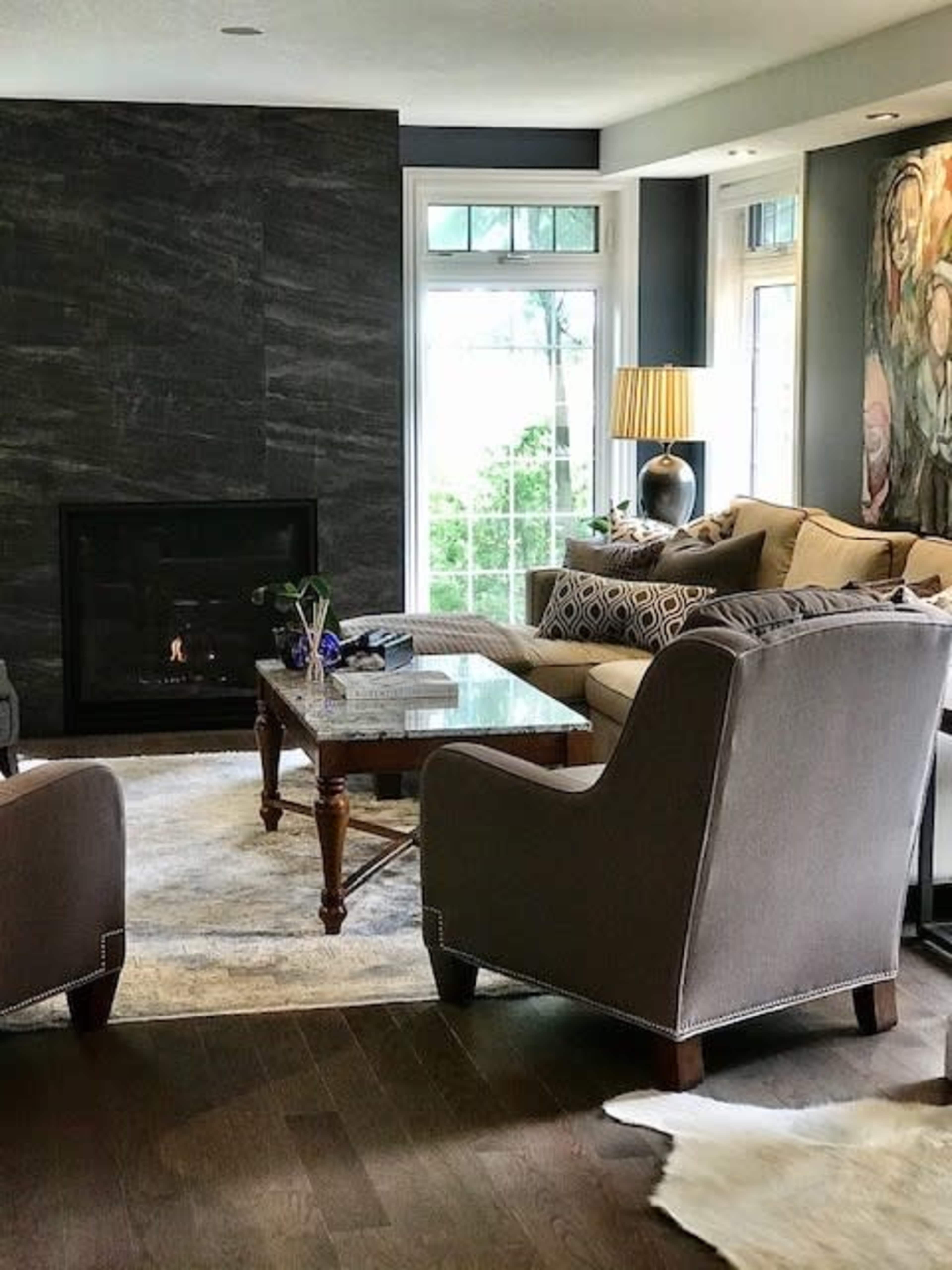 A modern living room with a gray stone accent wall, a cozy seating area featuring two armchairs and a sofa, a coffee table, and large windows allowing natural light.
