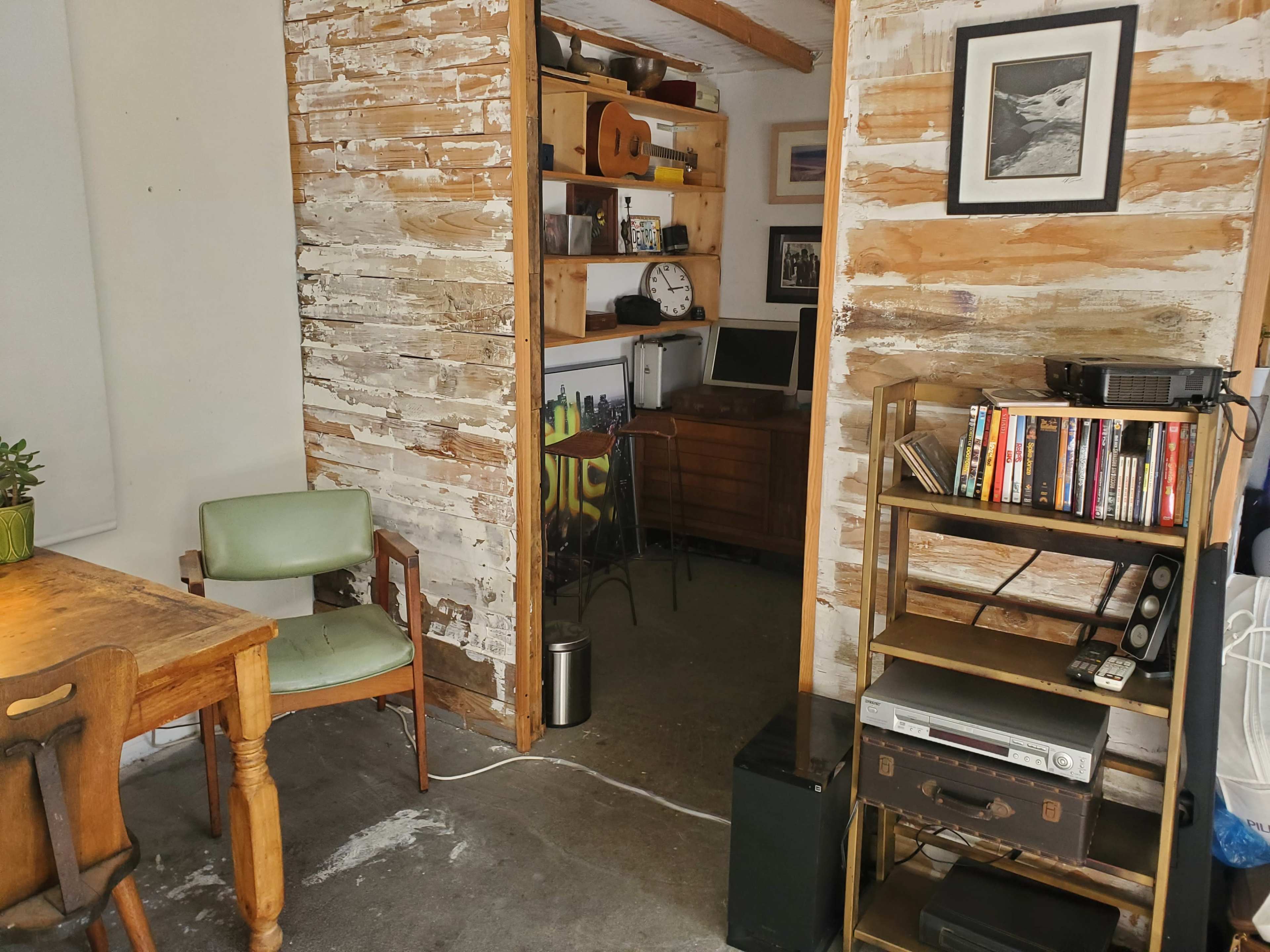The image shows a rustic room with exposed wooden walls, a small wooden table with a chair, a bookshelf filled with various items, and a computer setup in a nearby nook.