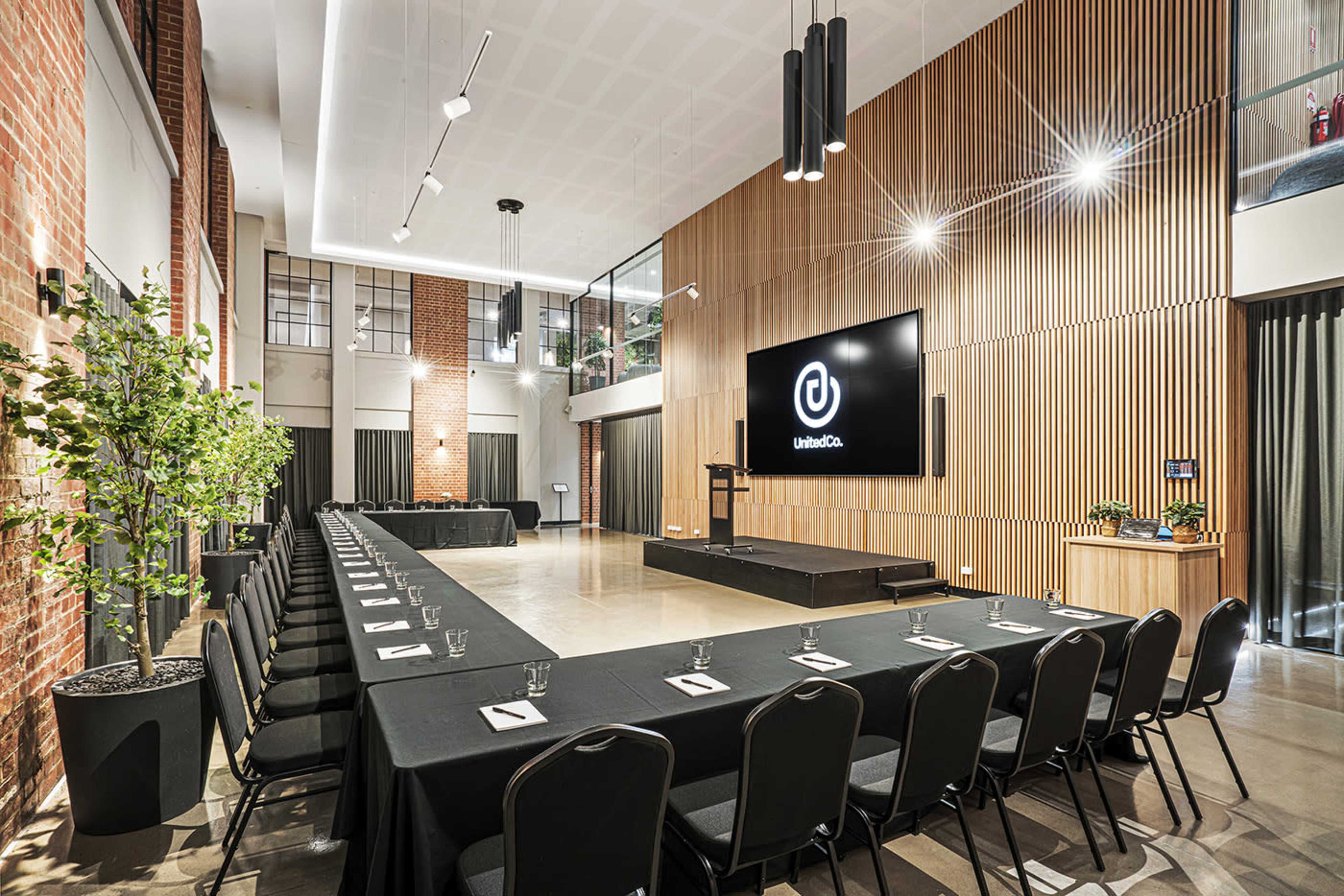 The image shows a spacious conference room with a long rectangular table set for a meeting, featuring a large screen on one wall and a podium at the front.