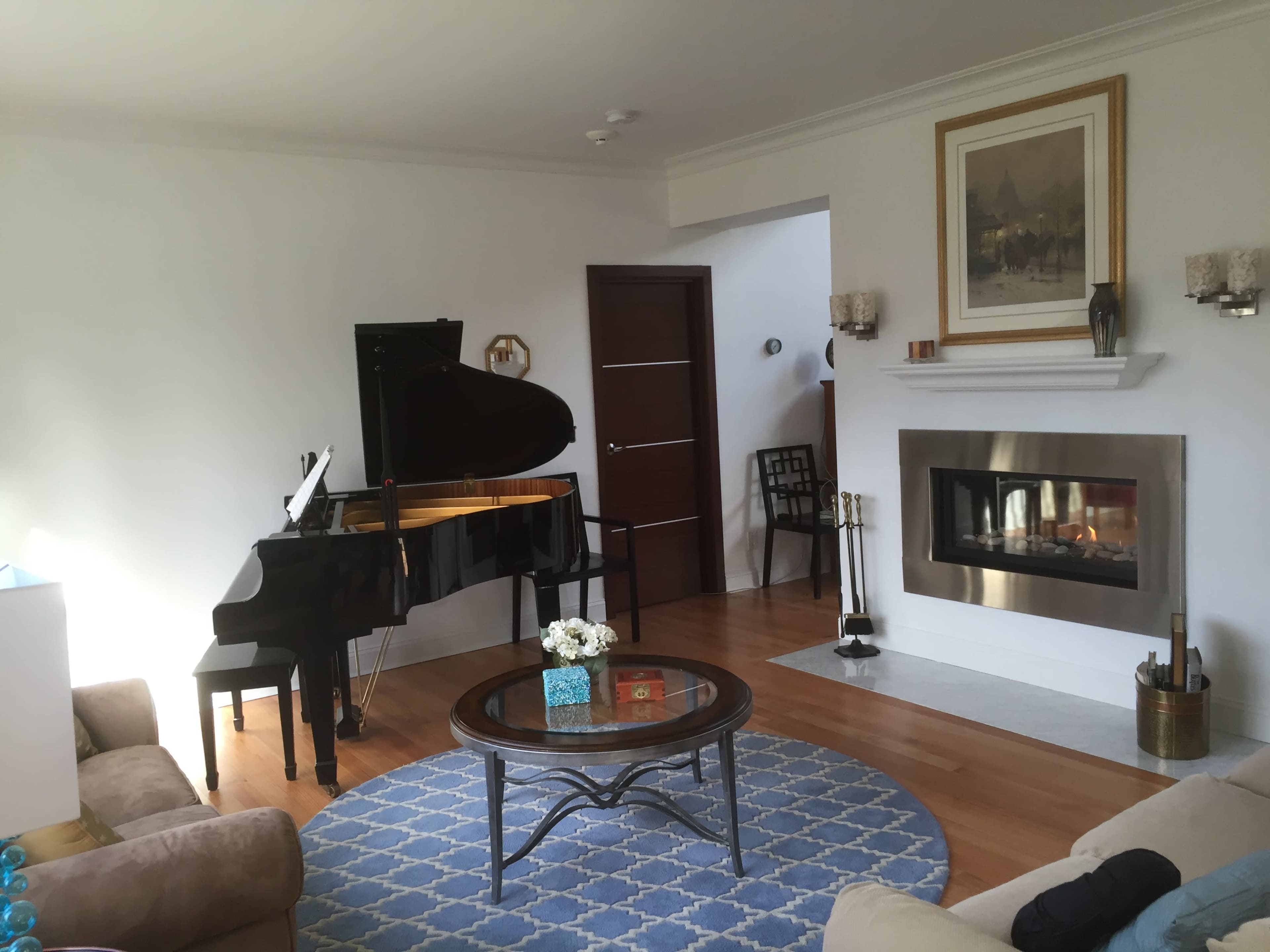 A modern living room features a black grand piano, a circular coffee table, and a fireplace with a framed artwork above.