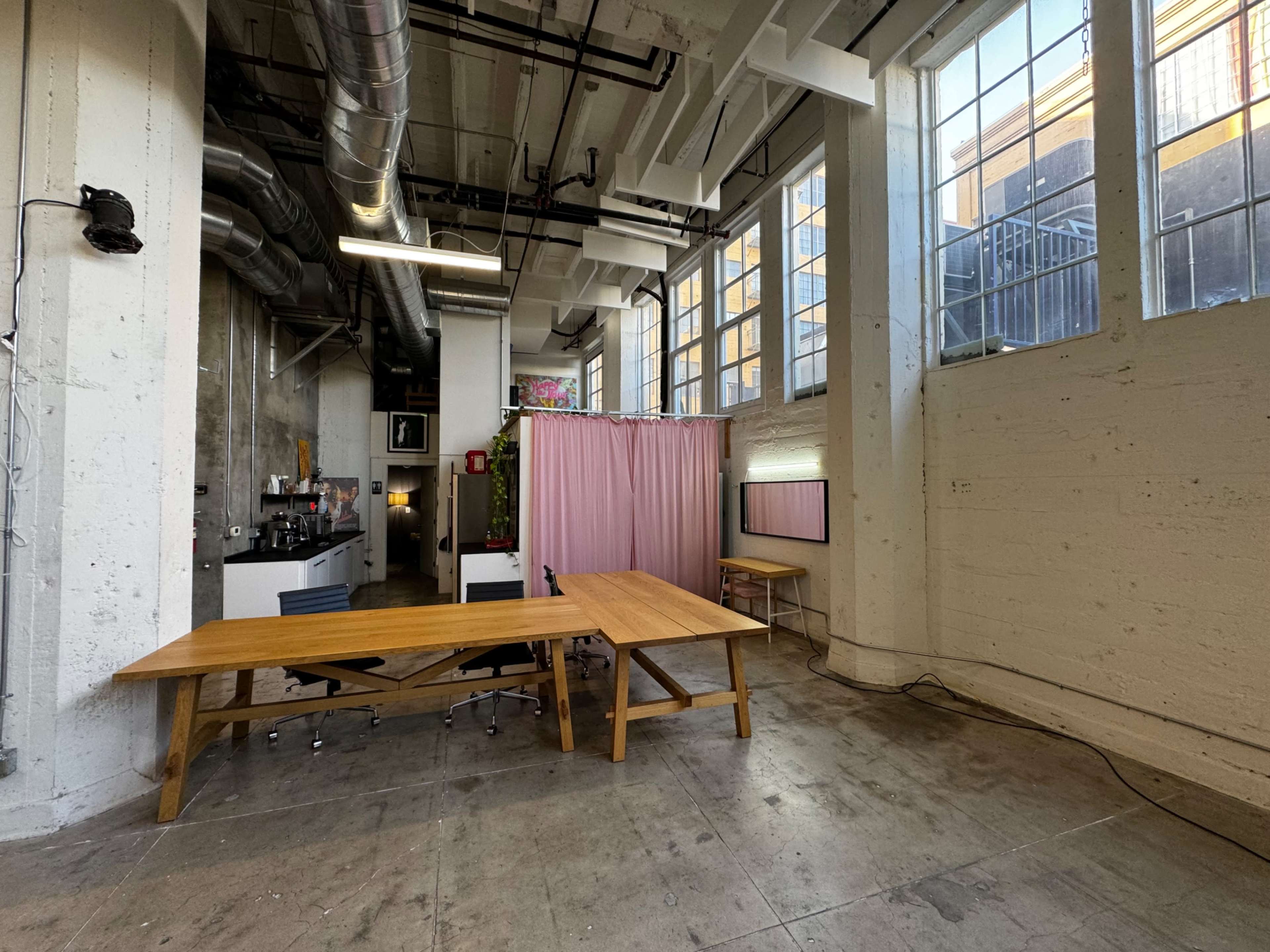 A spacious, industrial-style office with large windows, a wooden table in the foreground, and a pink curtain separating a section of the space.