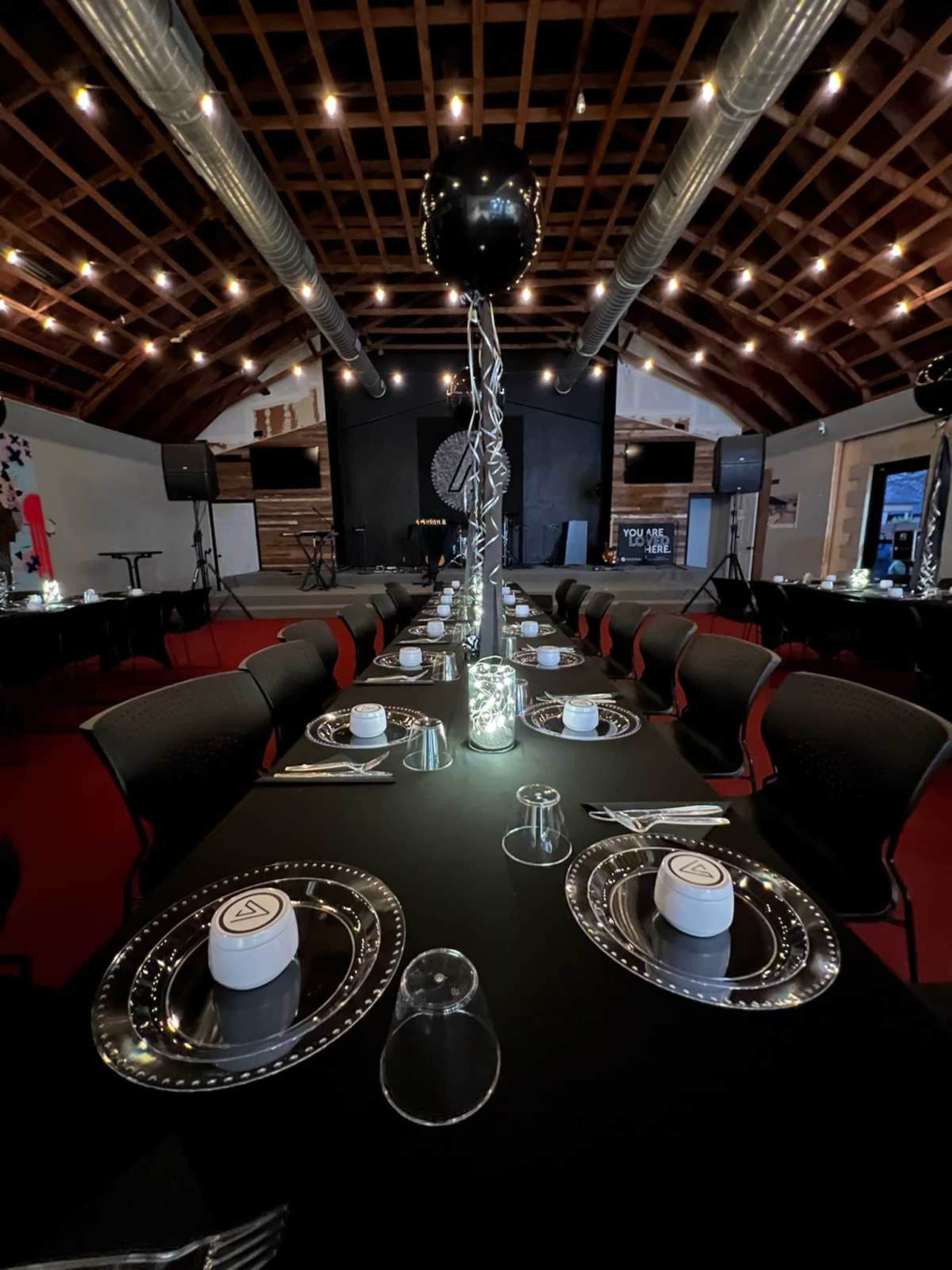 A long dining table is elegantly set with black tablecloths, metallic chargers, and centerpieces featuring silver balloons in a spacious venue.