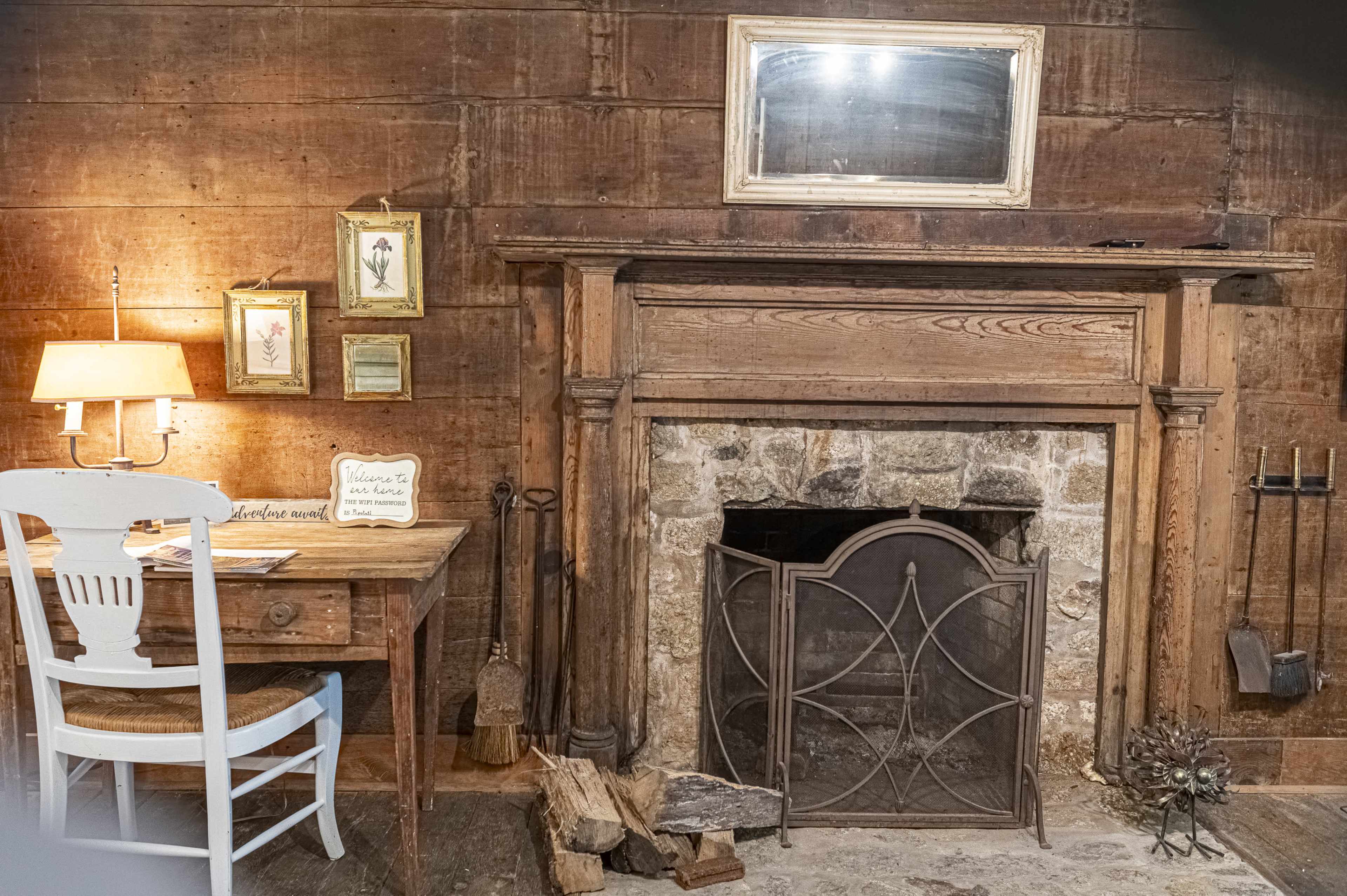 A rustic room with a wooden wall, a stone fireplace with a metal screen, a small table with a lamp, and framed pictures hanging nearby.