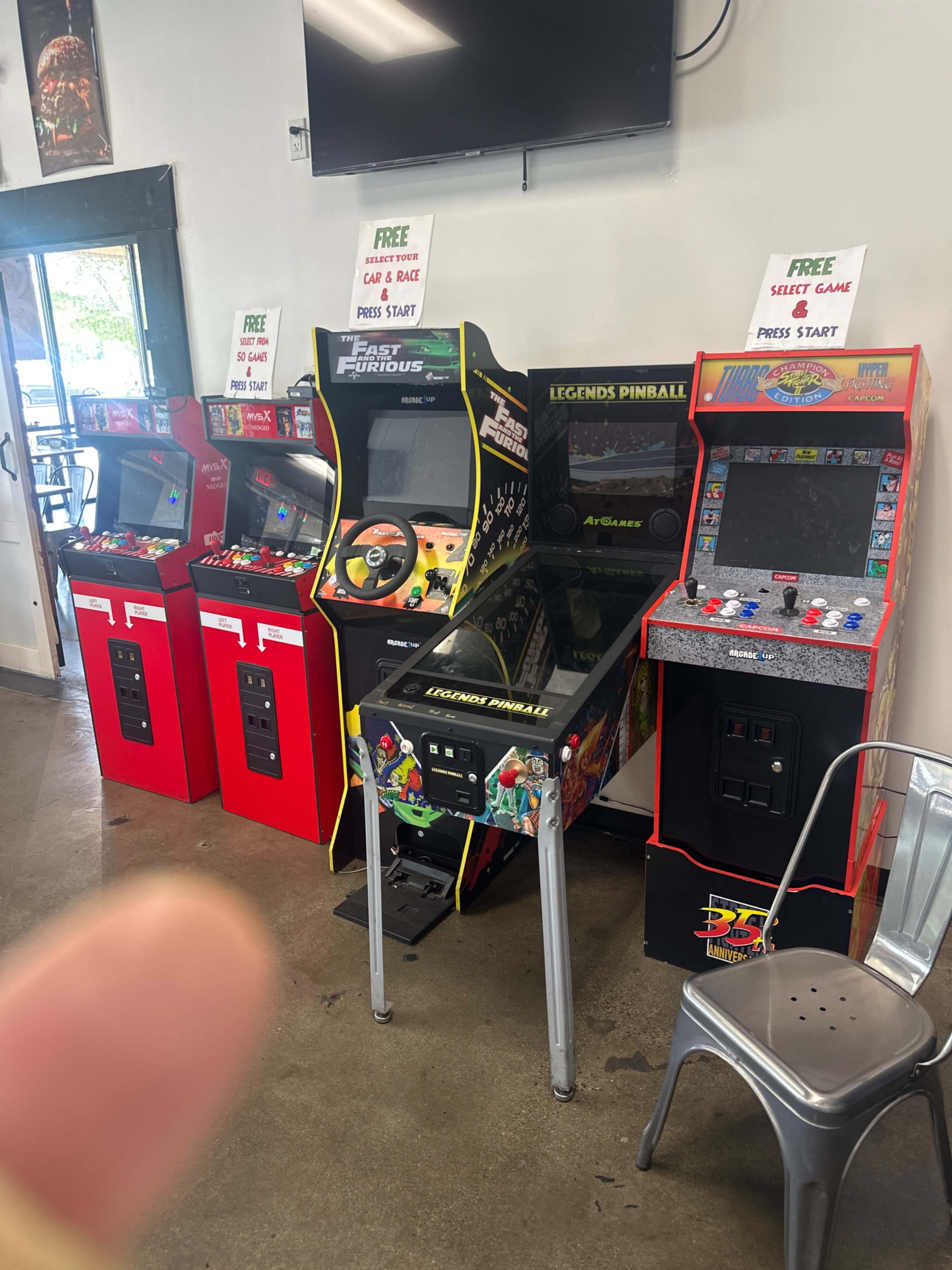 The image shows a row of arcade game machines, including a driving game, a pinball machine, and a classic arcade cabinet, set up in a recreational space.