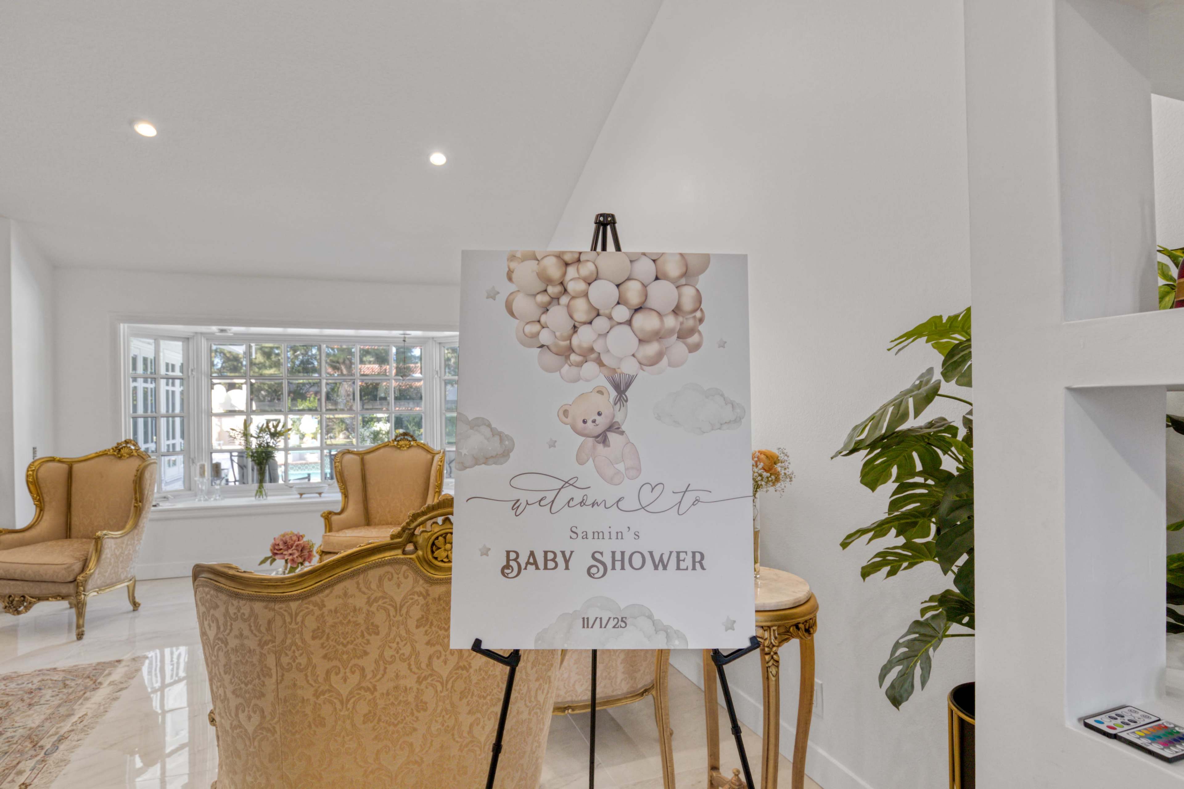 A decorative sign welcoming guests to a baby shower features a bear holding a cluster of balloons, set against a bright, airy room with elegant furniture.