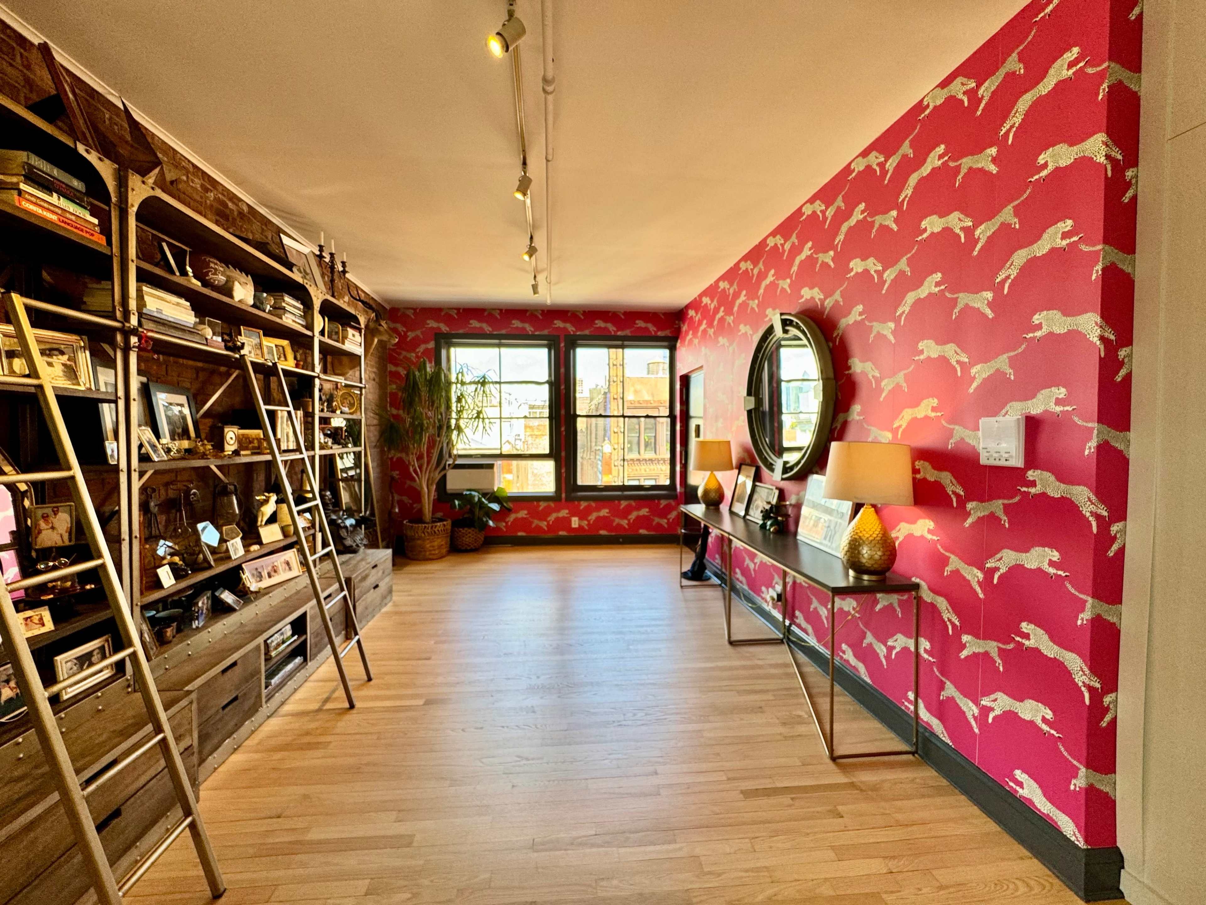 The image shows a brightly lit room with a pink wall featuring a pattern of white leopards, bookshelves on one side, and a console table with a round mirror on the opposite wall.