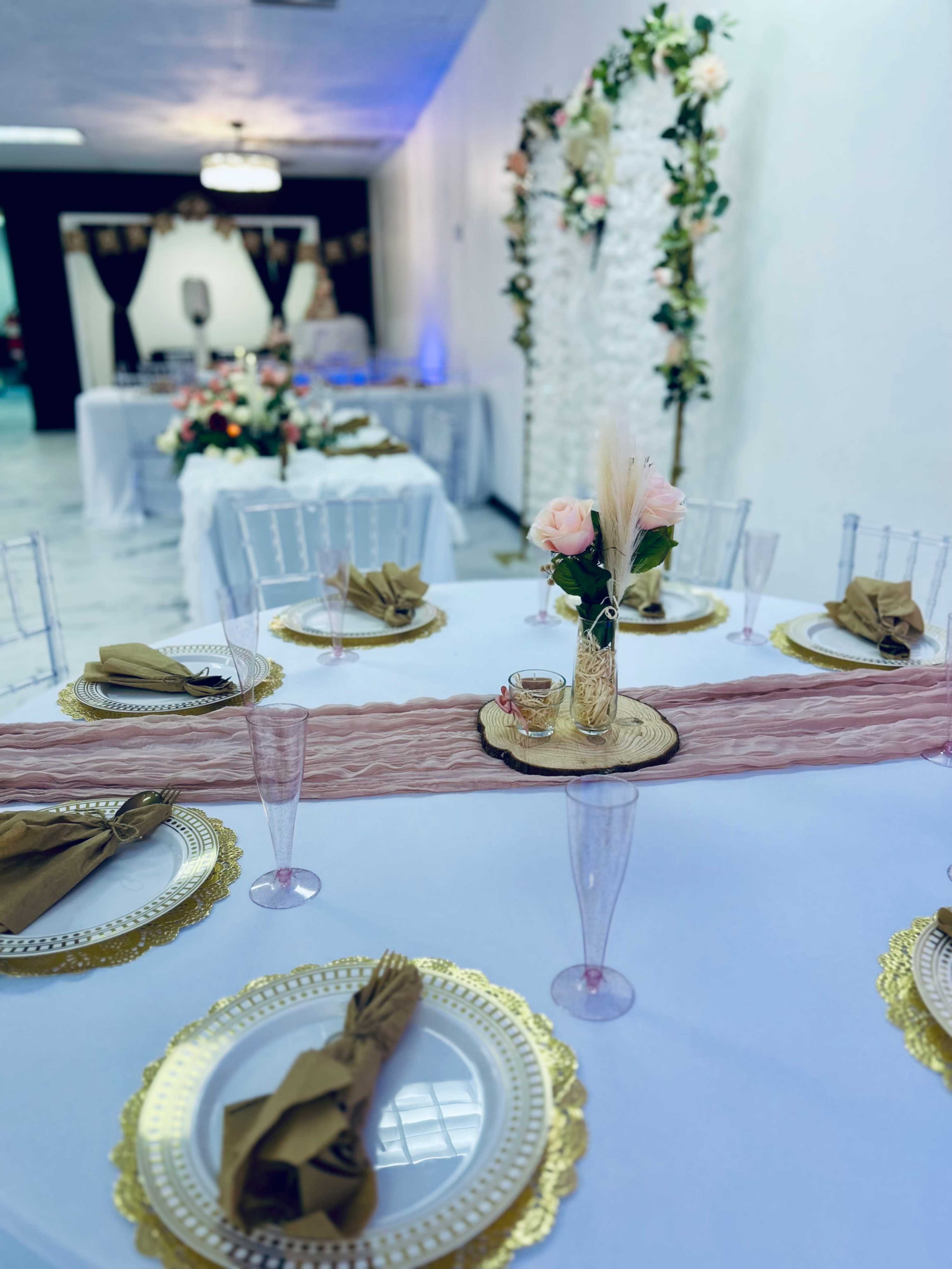 A decorated dining area features elegantly set tables with gold-rimmed plates, folded napkins, and floral centerpieces, against a backdrop of floral decorations on the wall.