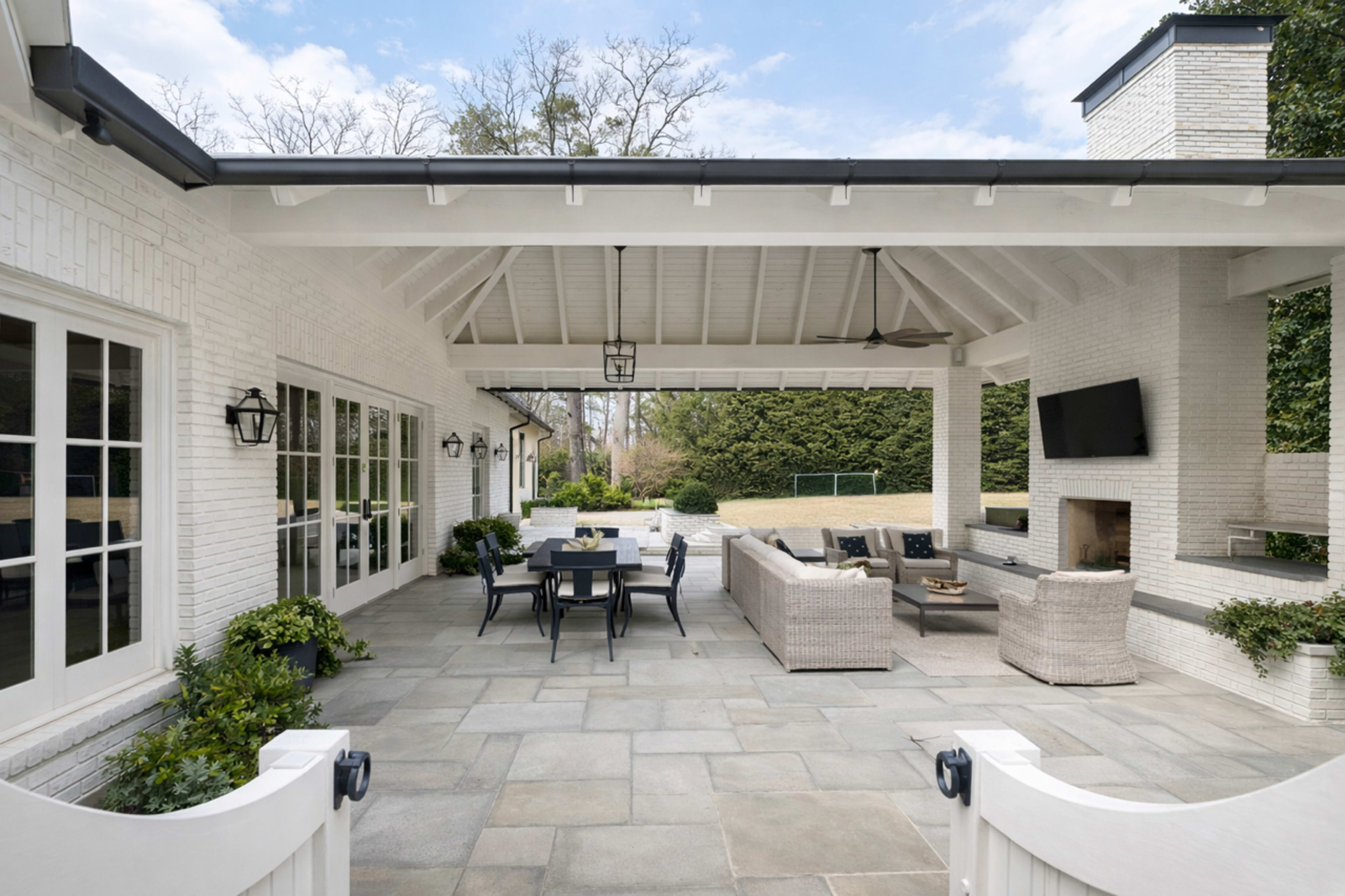 The image shows a spacious, covered outdoor patio area with a dining table, seating, and a fireplace, surrounded by greenery.