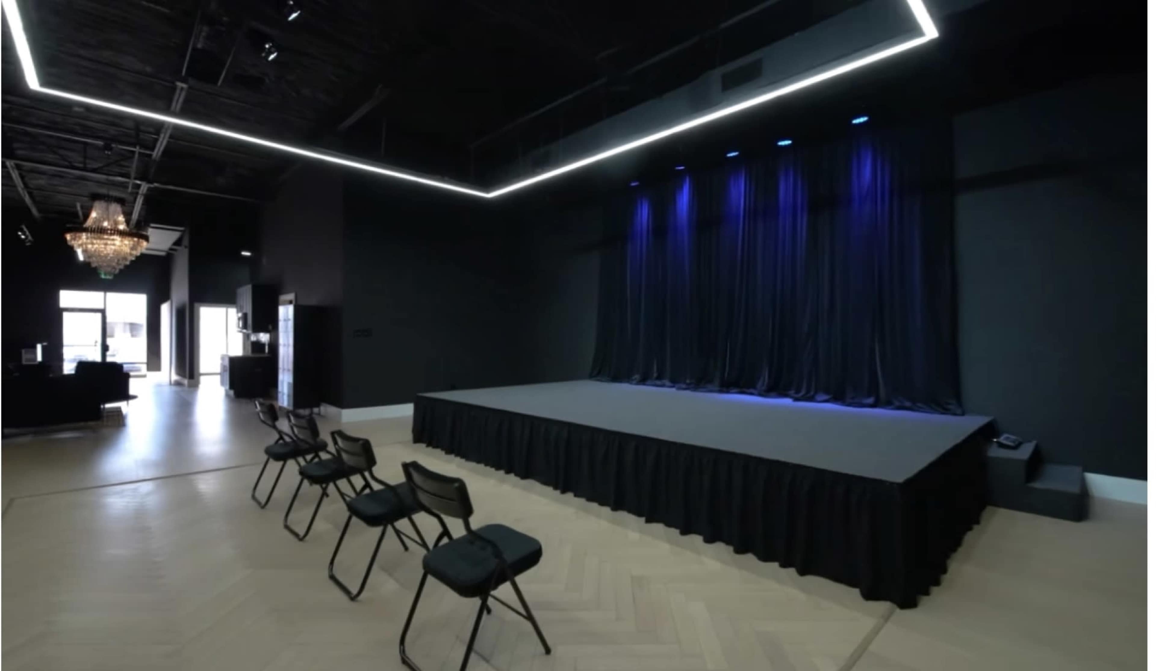 The image shows a modern performance space with a raised stage, black curtains, and a few empty chairs arranged in front of it.