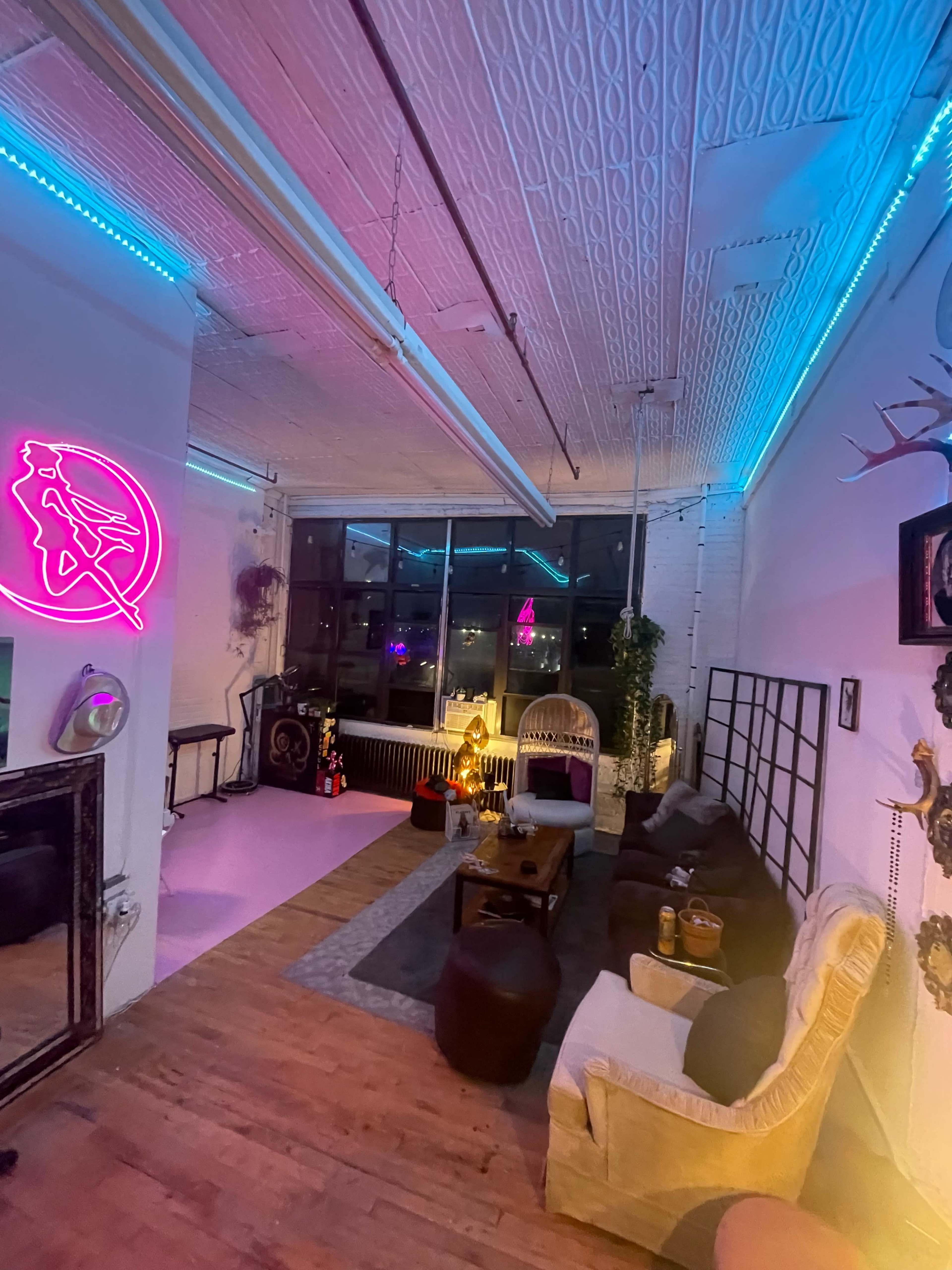 The image shows a stylish, modern lounge with eclectic furniture, bright neon lighting, and large windows.