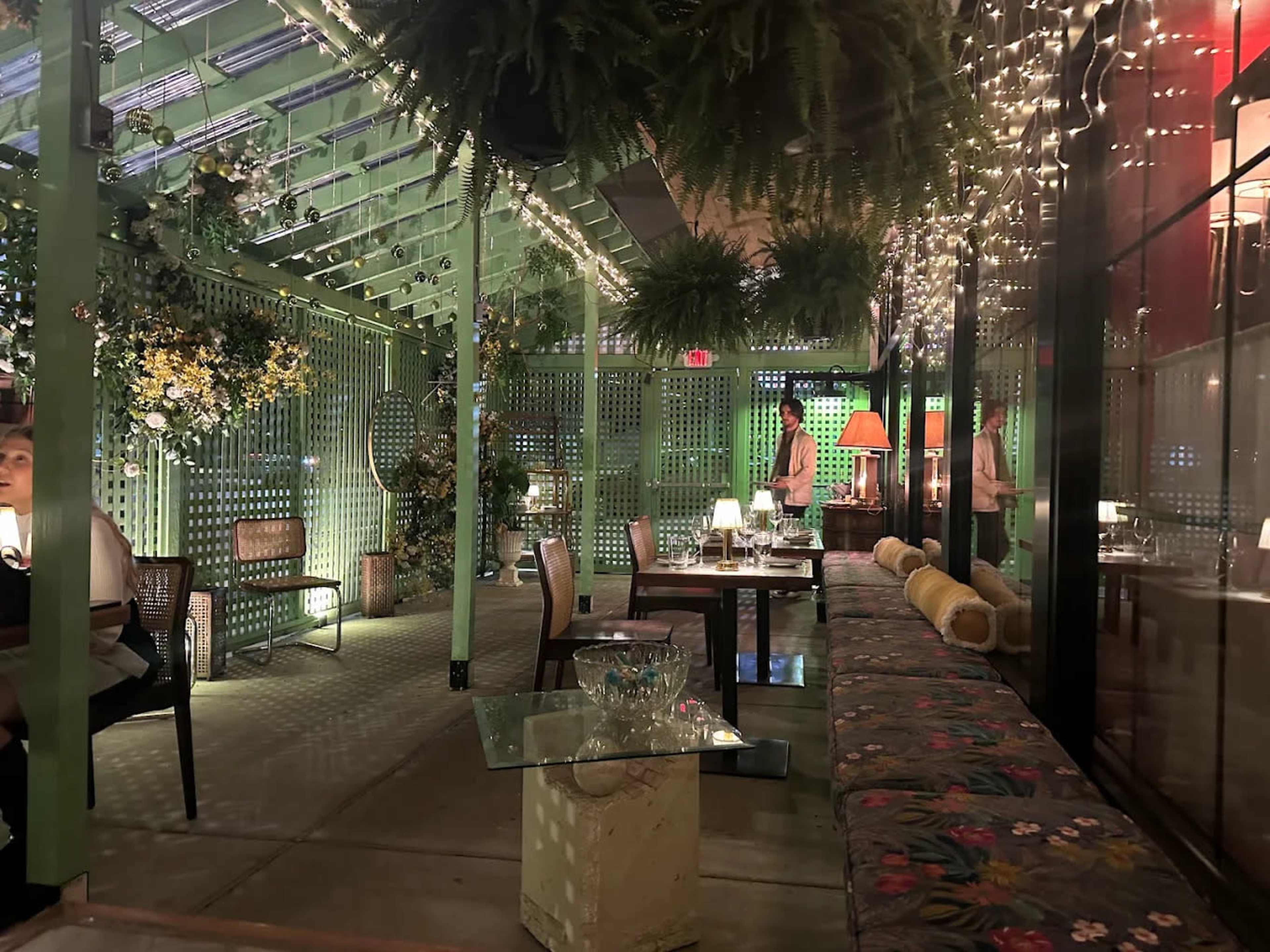 The image shows a vibrant indoor dining space with greenery, strings of lights, and wooden furniture arranged for patrons.
