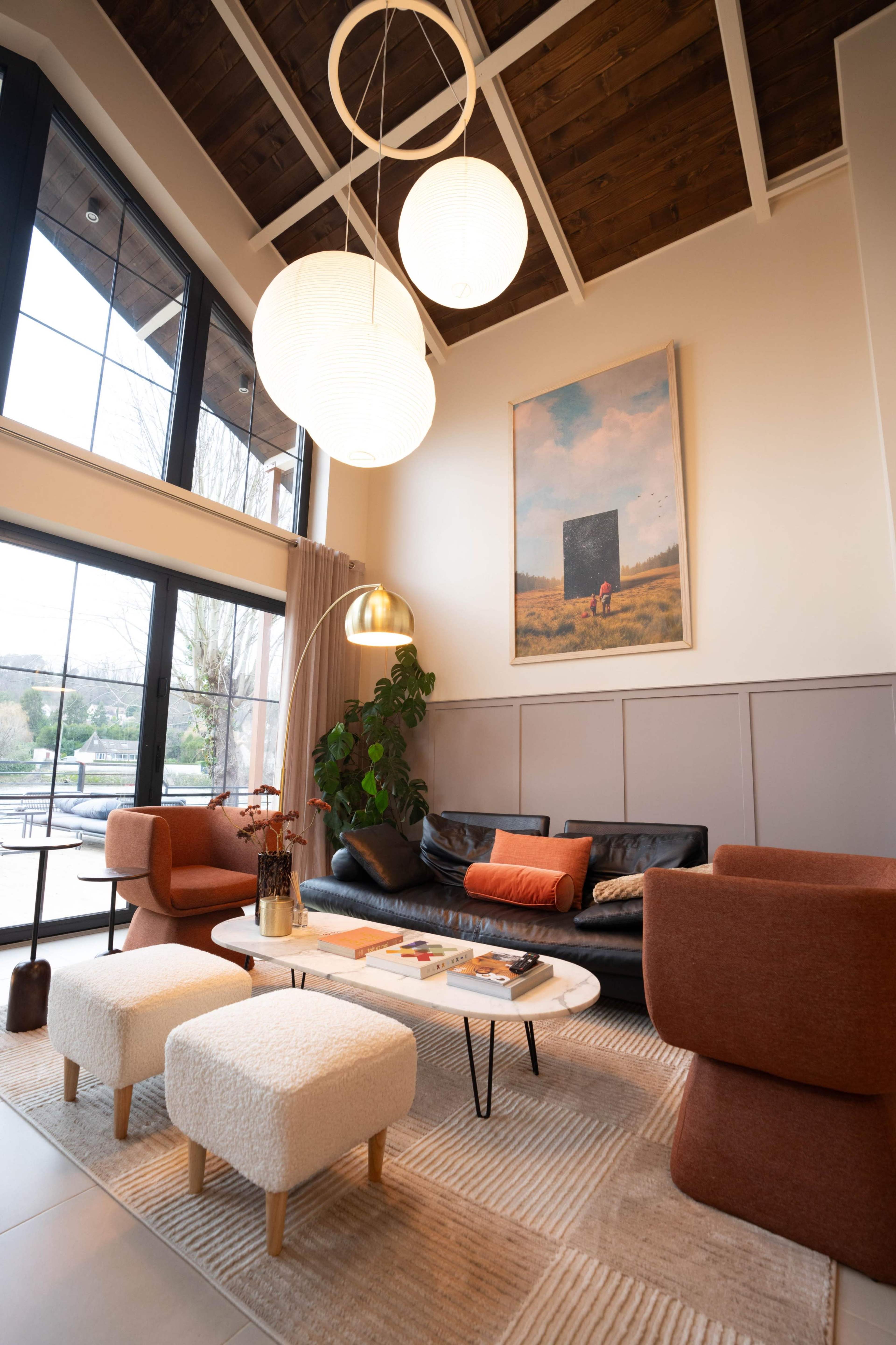 The image shows a modern living room featuring a black leather sofa, two orange accent chairs, a white coffee table, and large windows with natural light.