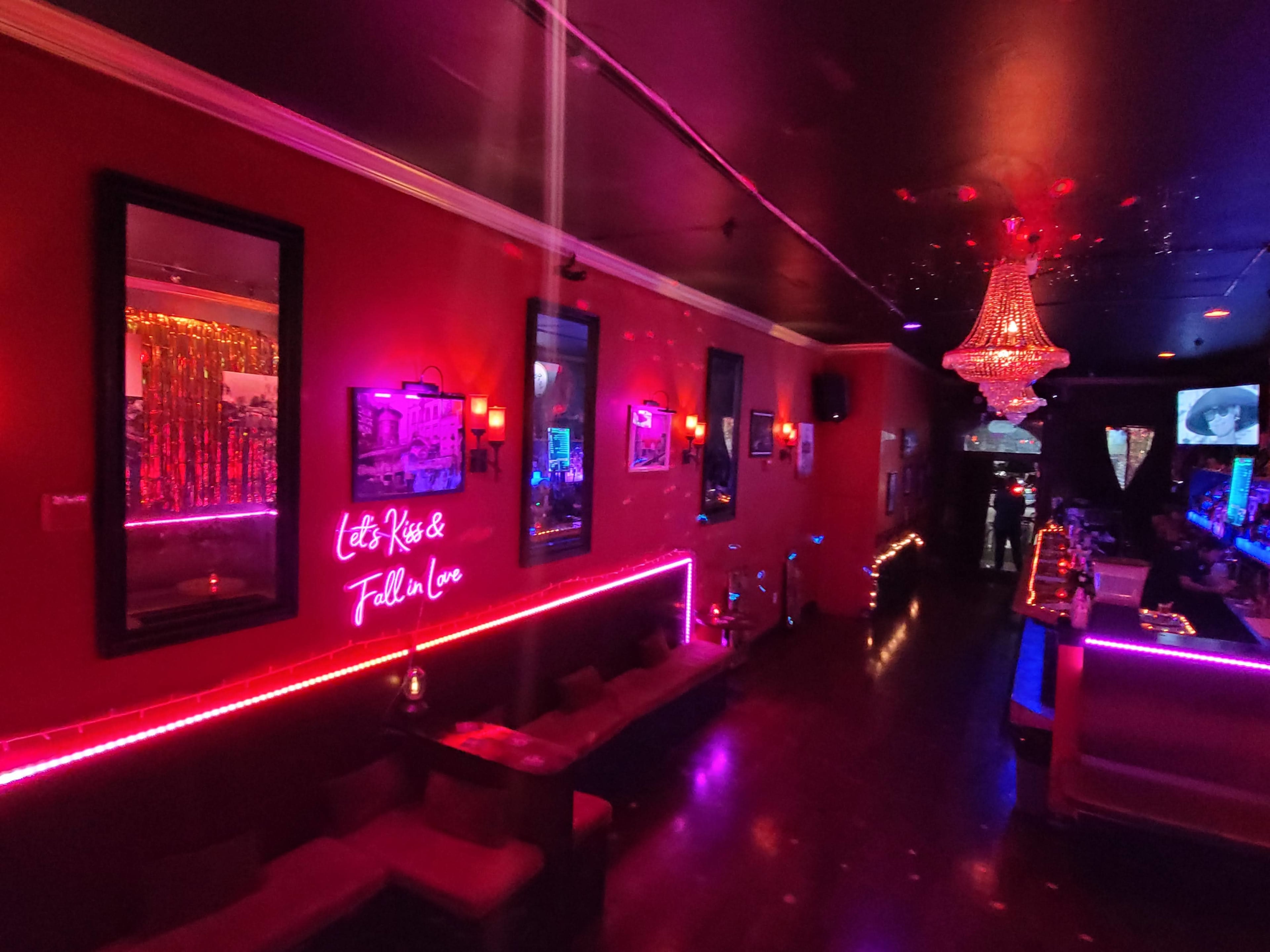 A dimly lit bar with neon decorations, mirrors on the walls, and a central chandelier illuminating the space.