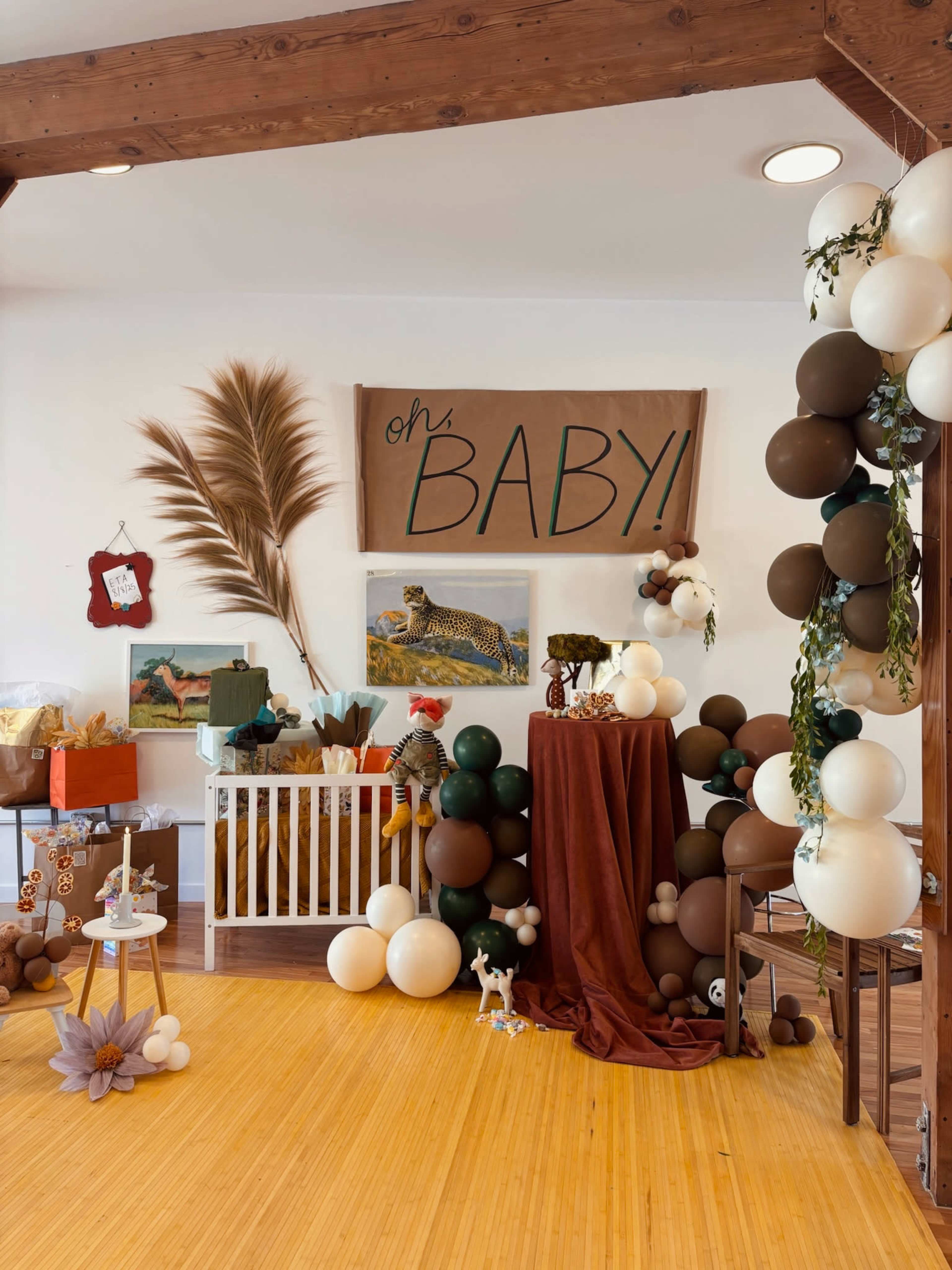The image shows a cozy baby shower setup featuring a decorated area with balloons, a display table, and themed artwork on the walls.