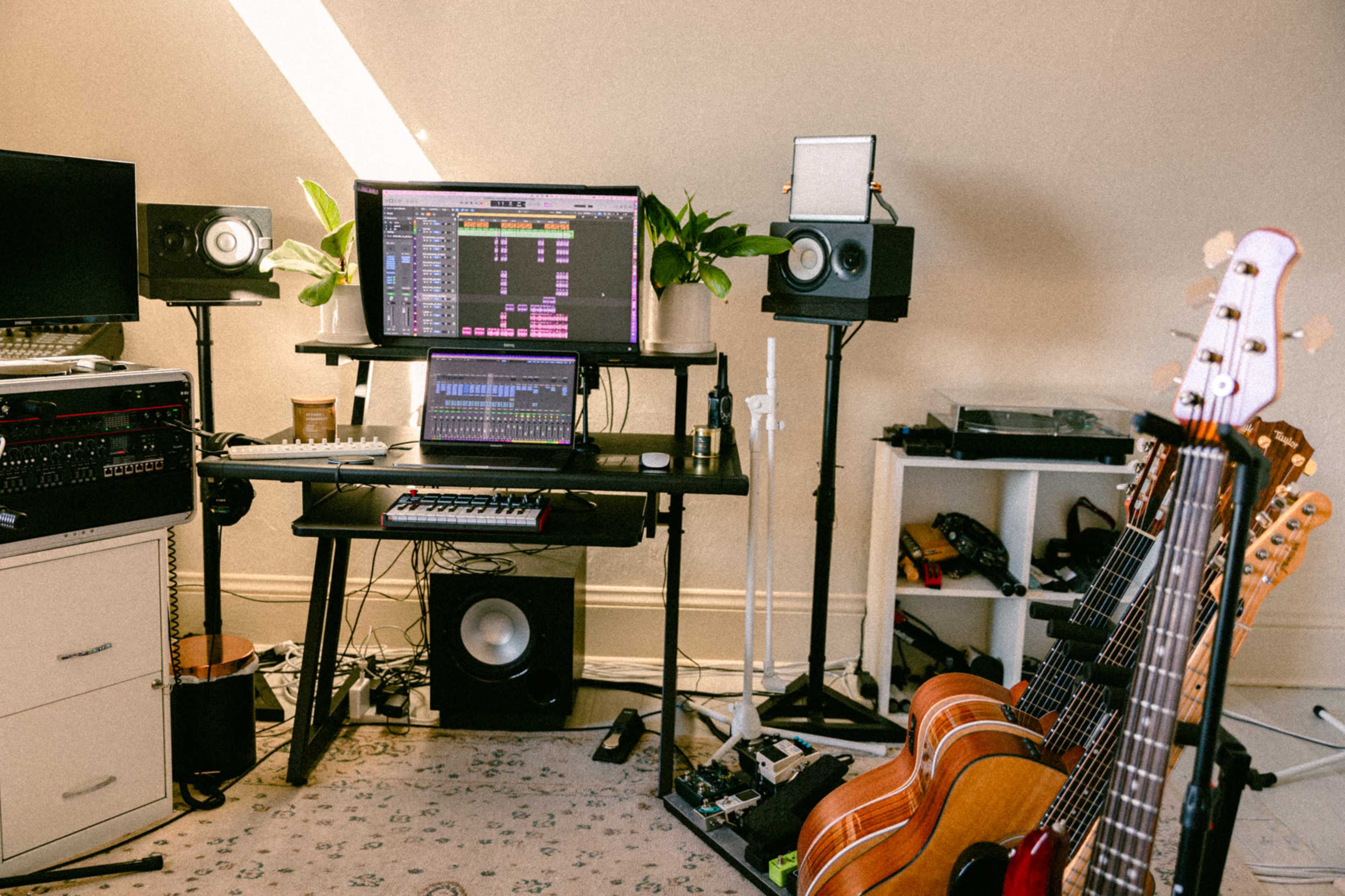 A home music studio features a desk with multiple monitors displaying audio software, speakers on stands, and a collection of guitars leaning against the wall.