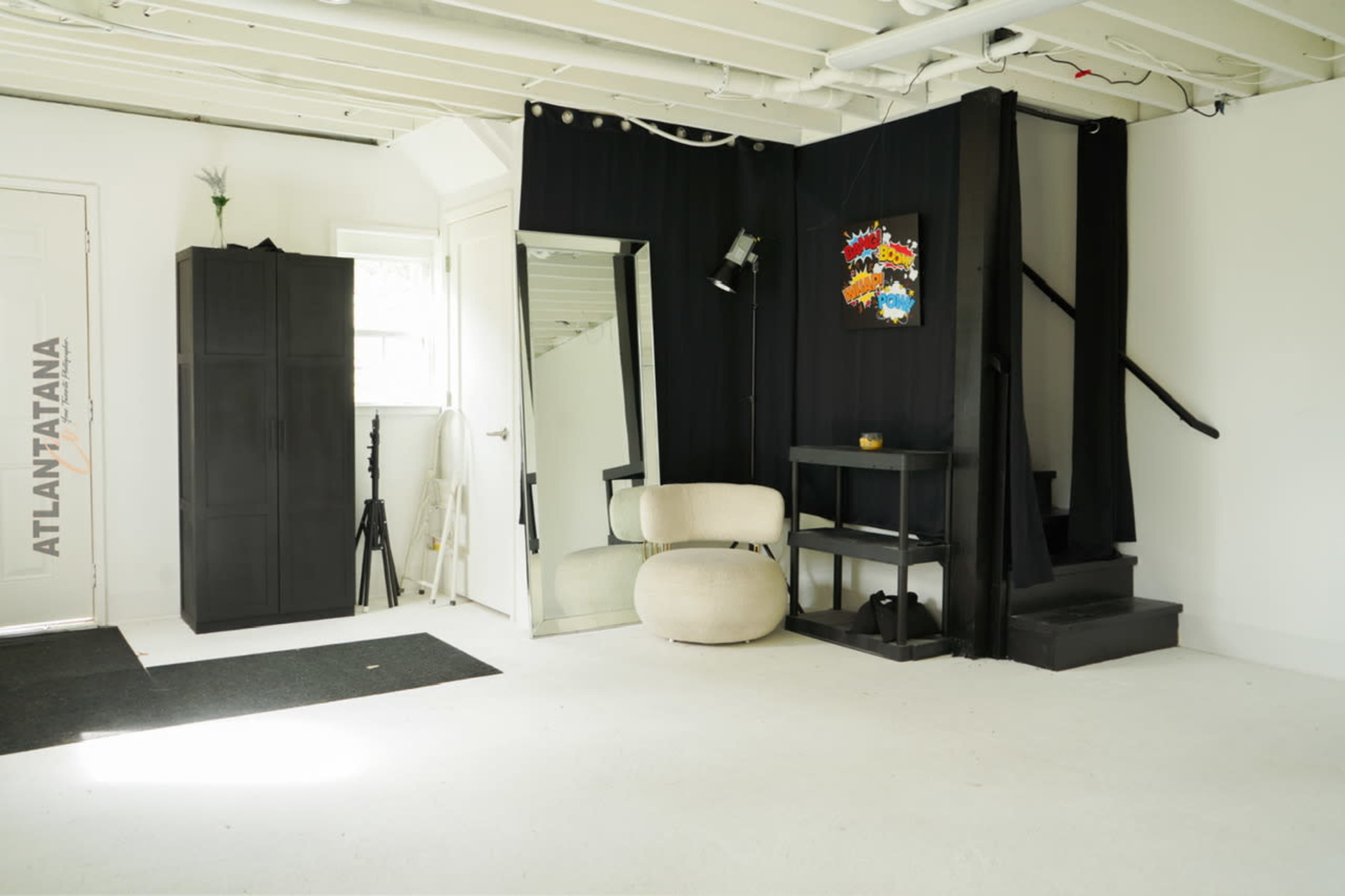 The image shows a minimalistic basement space featuring black furniture, a large mirror, and a staircase leading to an upper level.