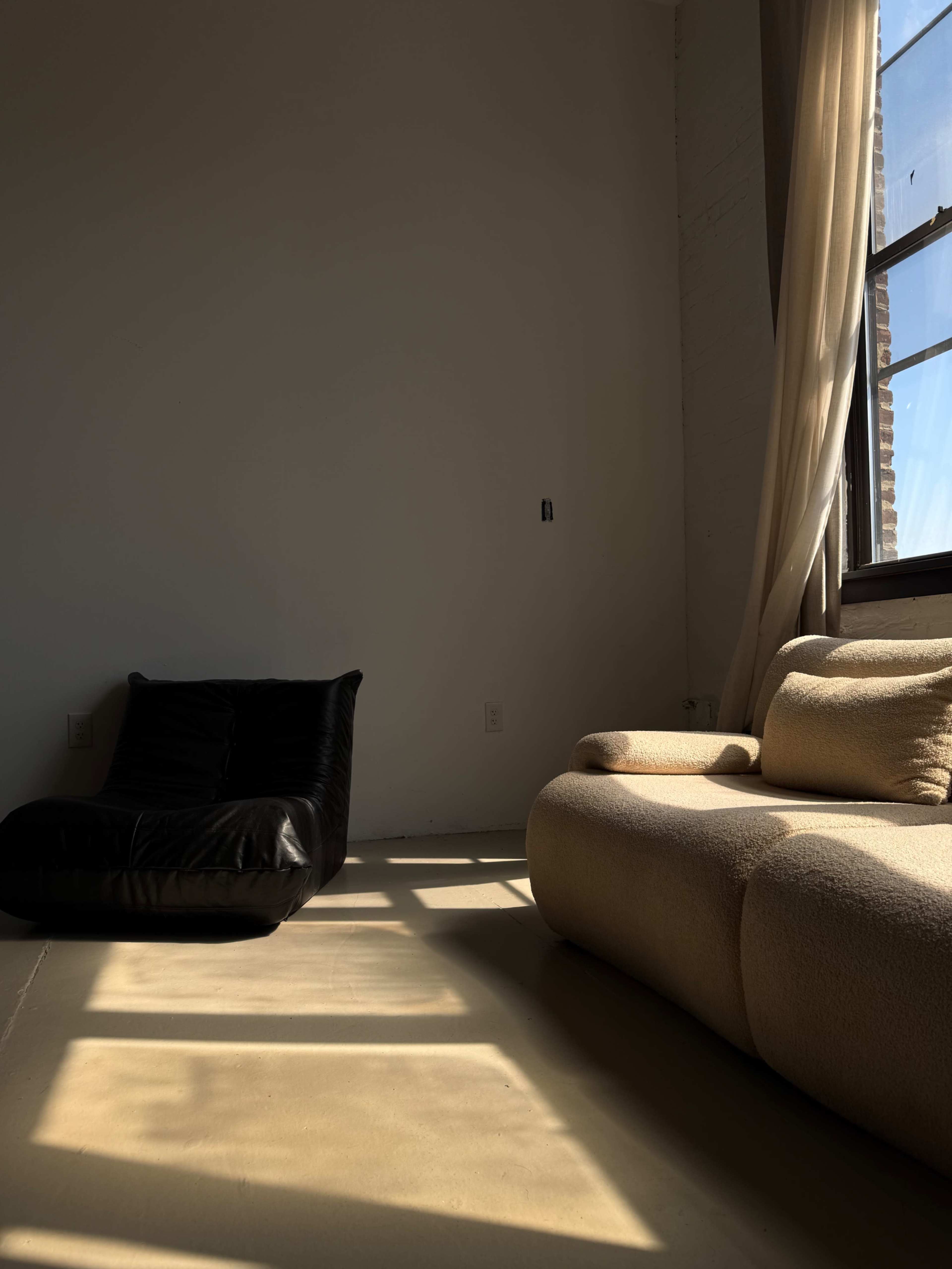 The image shows a minimalist interior with a black bean bag chair positioned next to a cream-colored couch, illuminated by sunlight streaming through a large window.