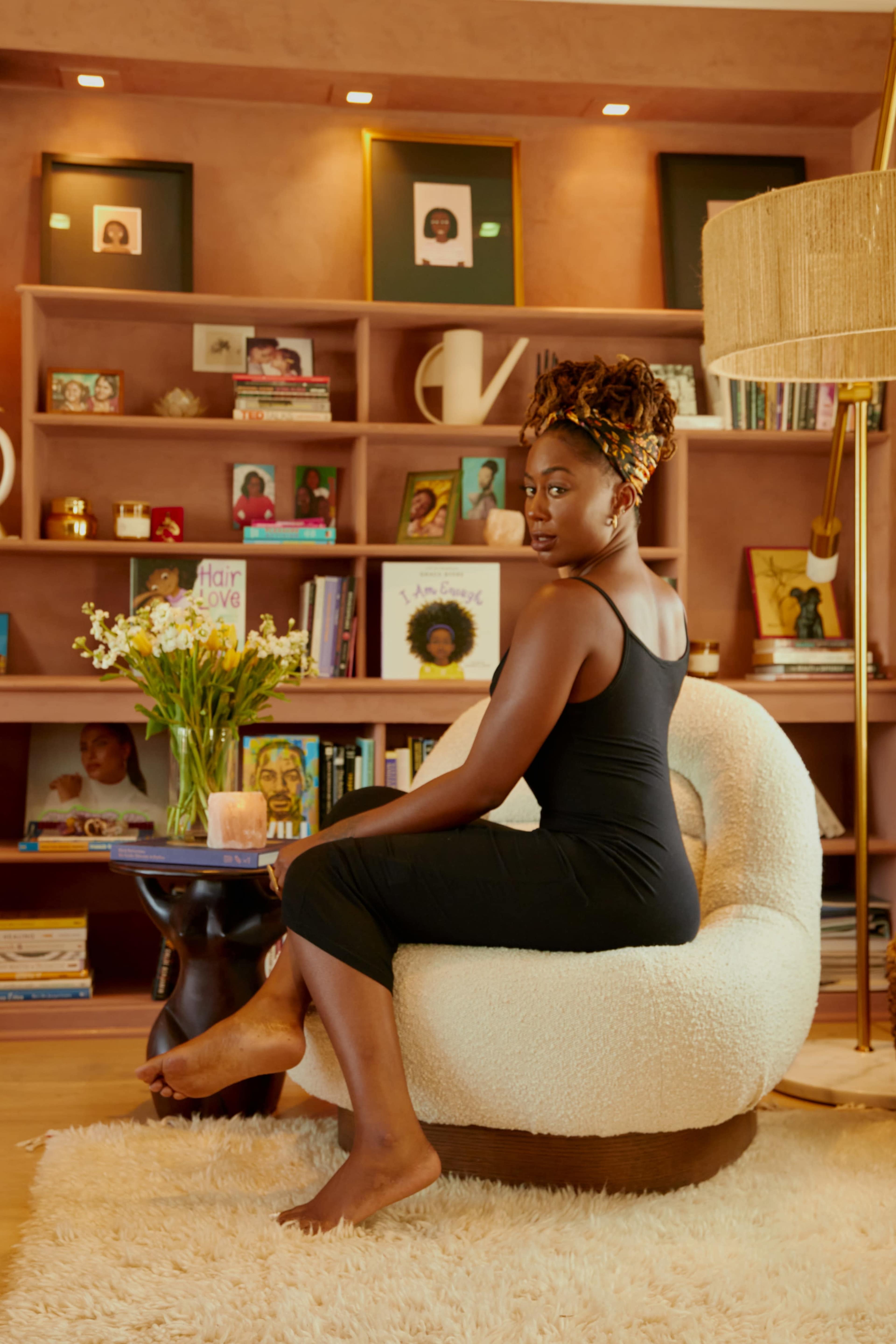 A woman sits in a cozy armchair in a well-decorated room with shelves displaying books and framed photographs.
