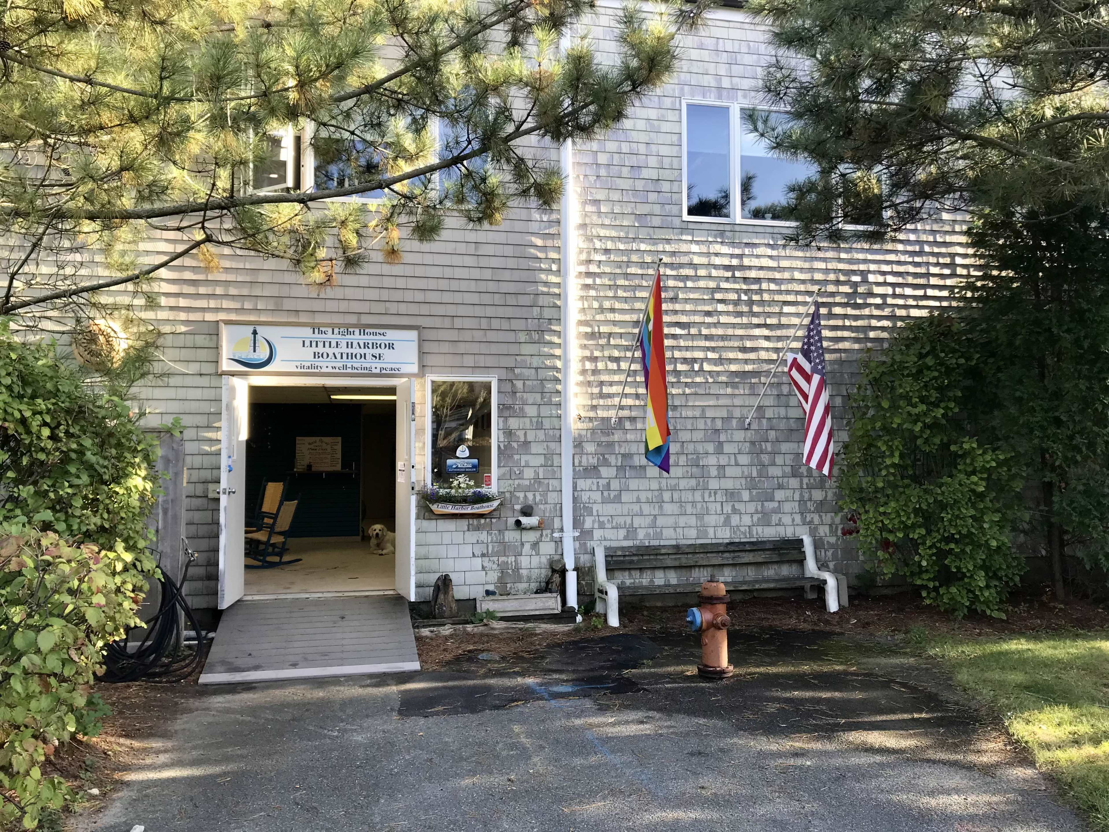 A building entrance with a sign for "Little Harbor Education," featuring a rainbow flag and an American flag, alongside a bench and a visible dog.