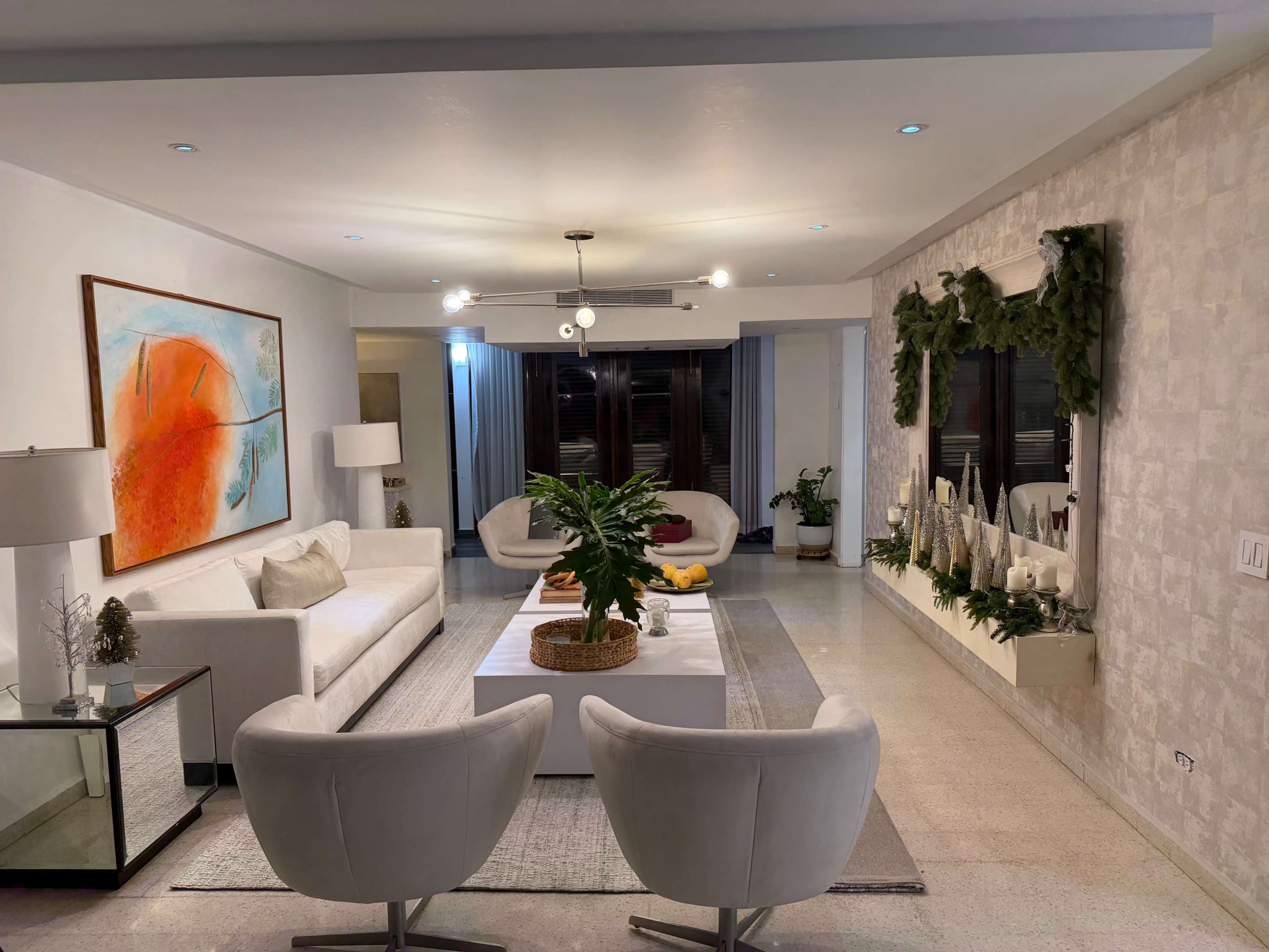 The image shows a modern living room featuring a white sofa, two light-colored chairs, a coffee table, and decorative plants, with artwork and holiday decorations adorning the walls.