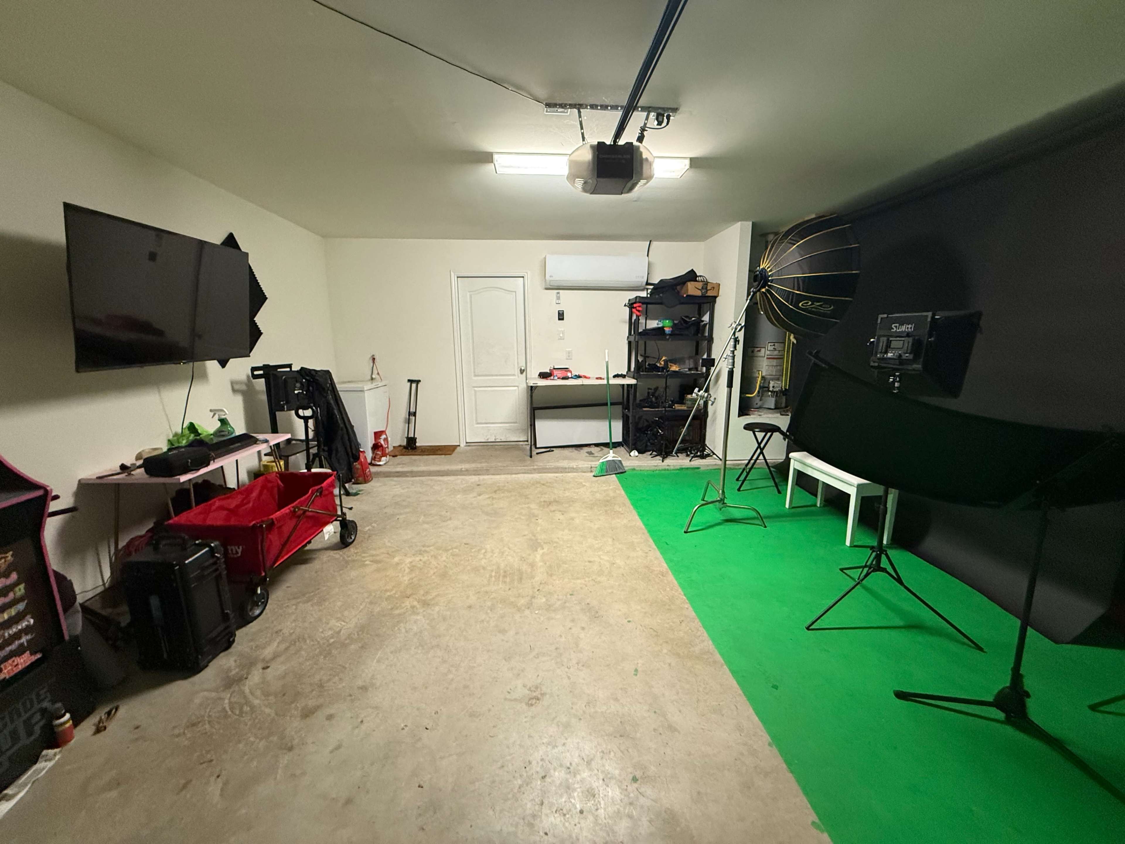 The image shows a garage space with a television on the left, storage shelves, a green backdrop, and a light setup for photography on the right.