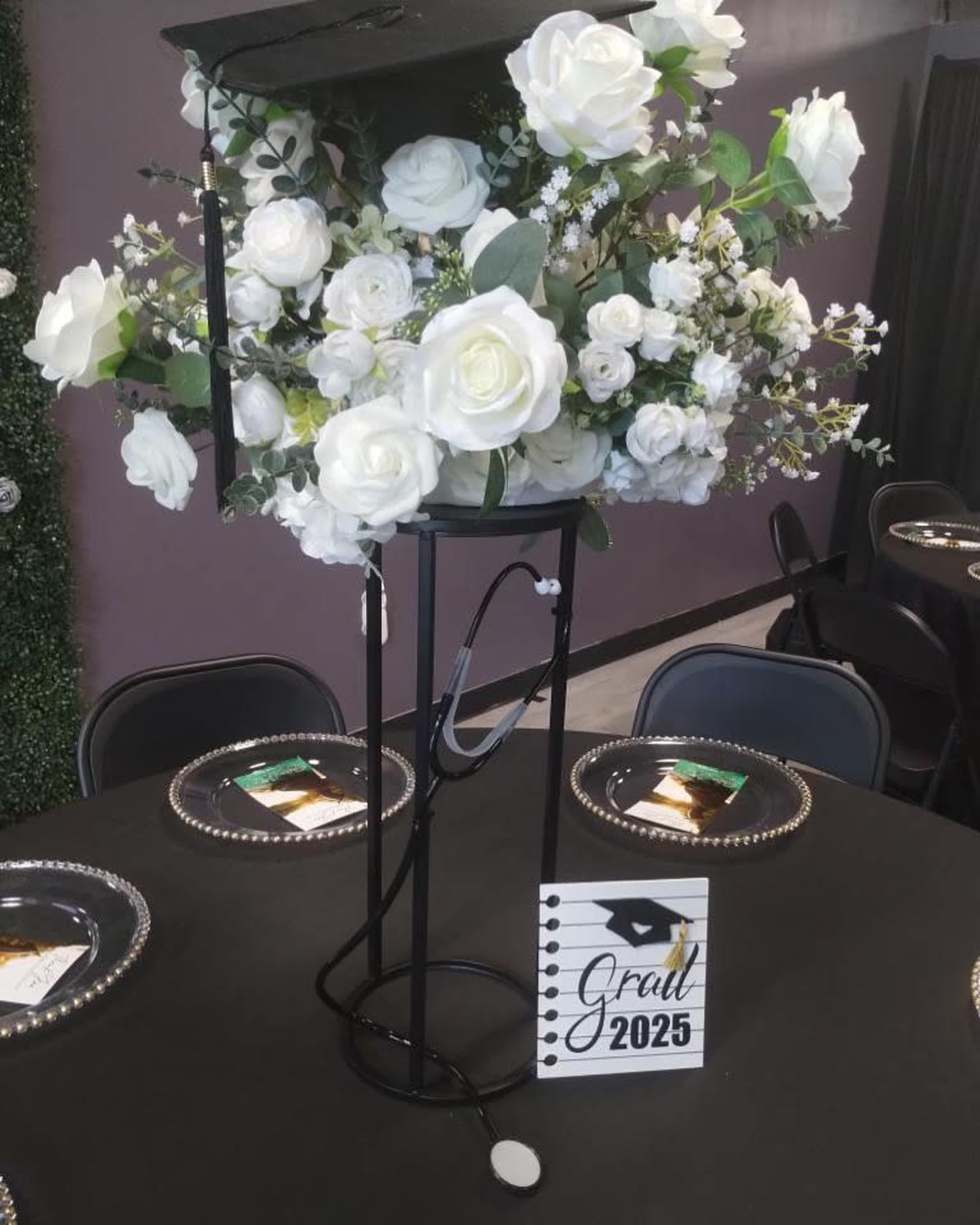 The image shows a table centerpiece featuring a graduation cap atop a floral arrangement of white roses and greenery, with a sign reading "Grad 2025" beside it.