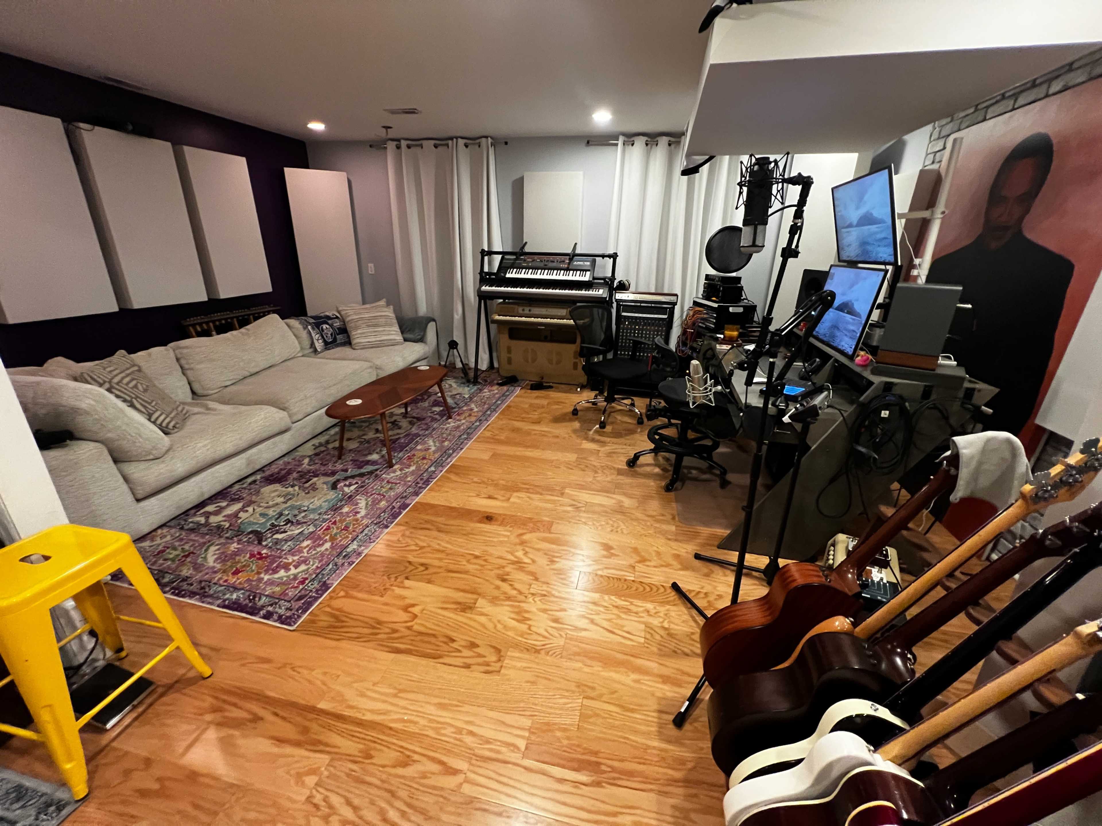 The image shows a cozy home studio with a sectional couch, a coffee table, musical instruments, and recording equipment arranged in a well-lit space.
