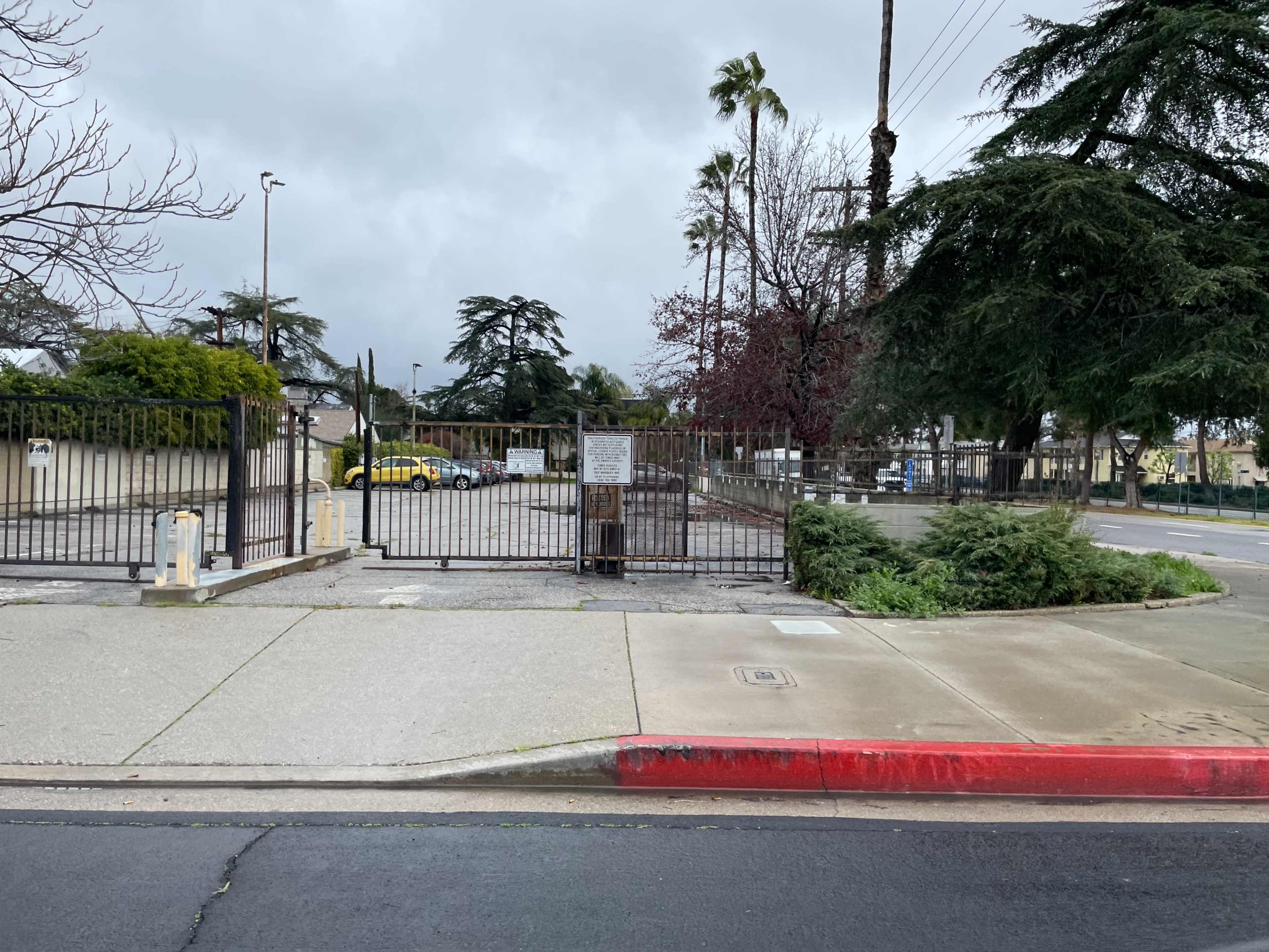 The image shows a fenced area with a gated entrance, surrounded by trees and shrubbery under a cloudy sky.