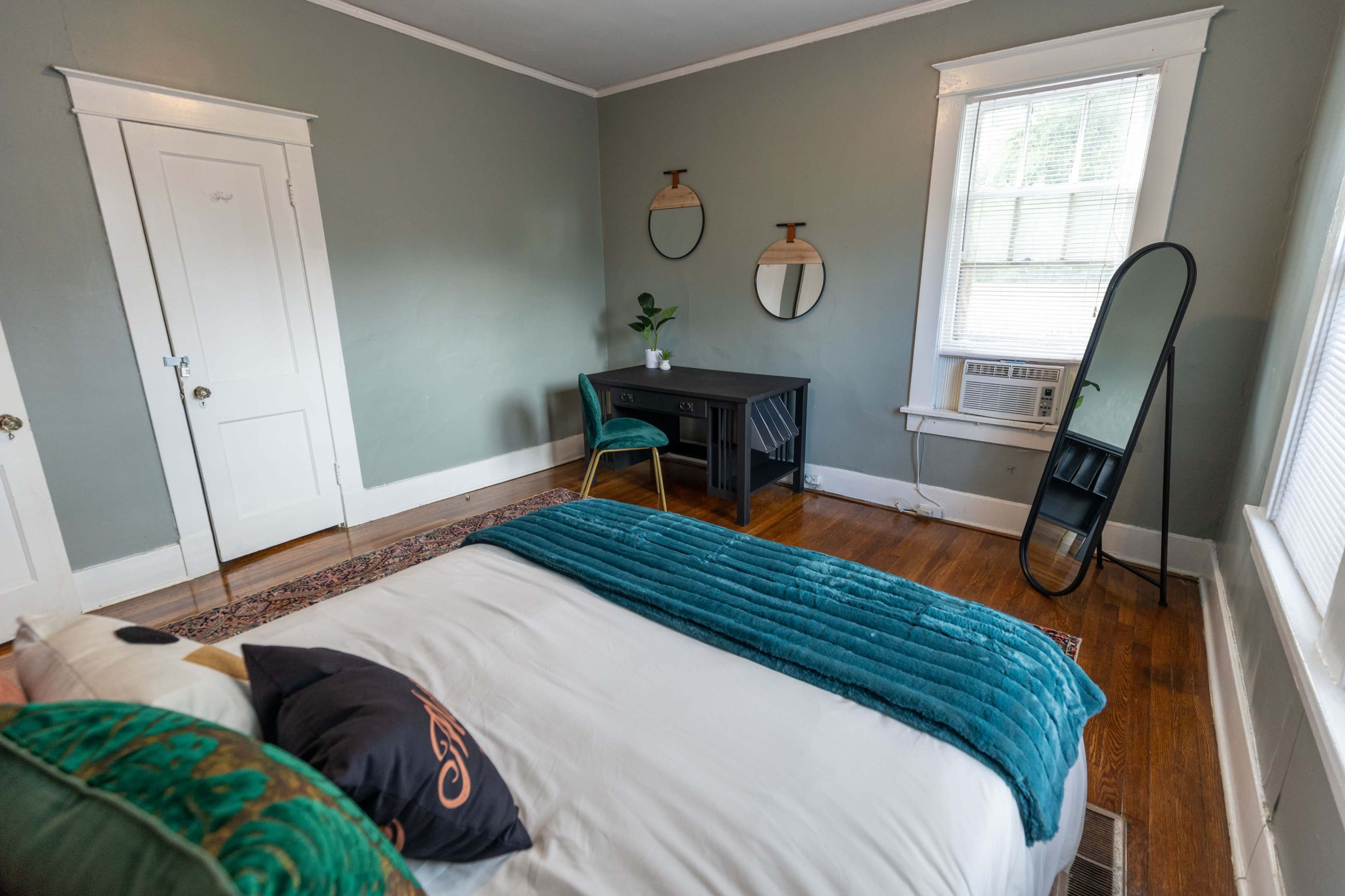 A cozy bedroom with a bed, a small desk with a chair, a mirror, and an air conditioning unit, all set against a light green wall.