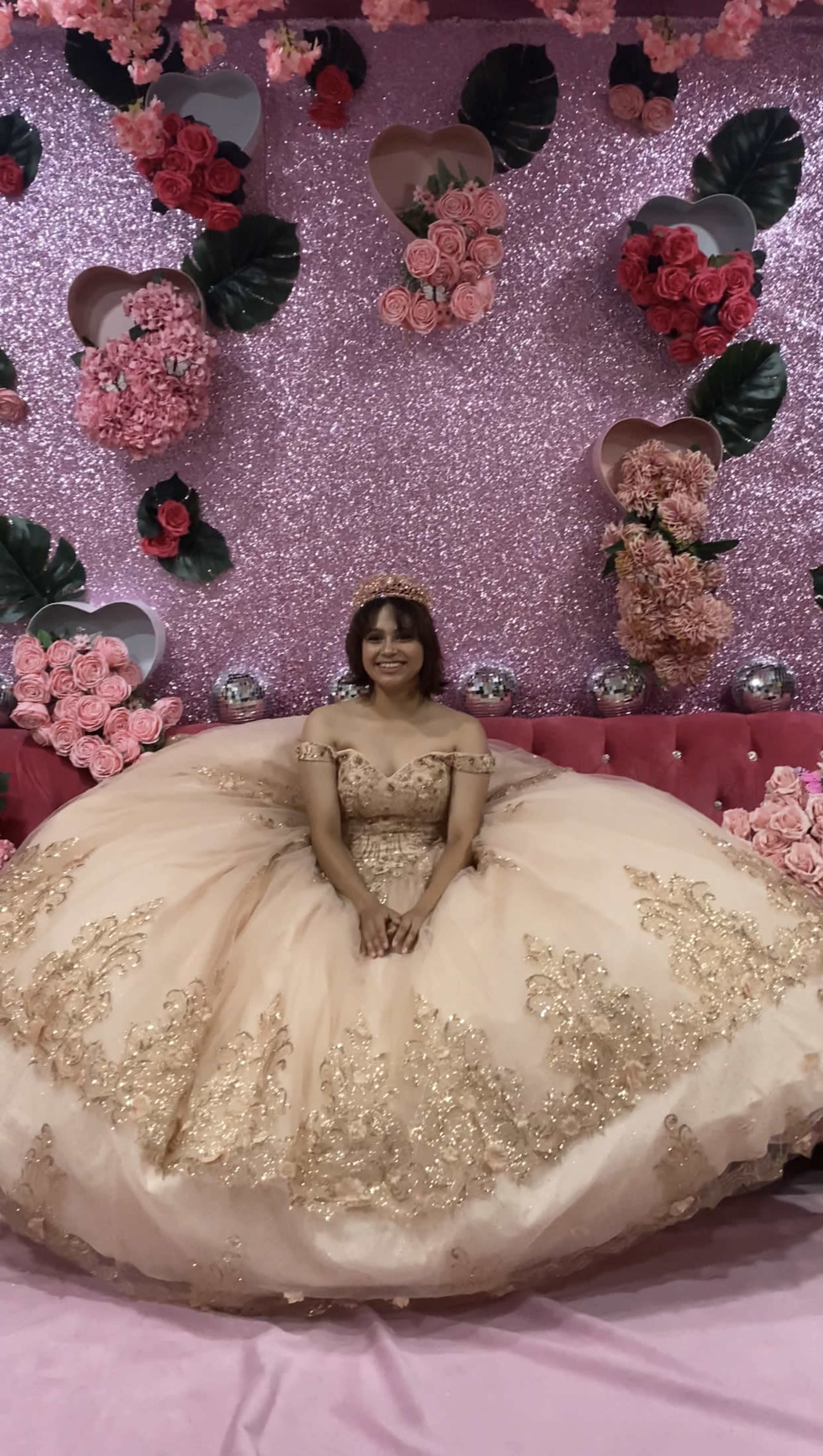 A girl in a golden embellished dress sits on a pink sofa surrounded by a sparkling pink backdrop adorned with flowers.