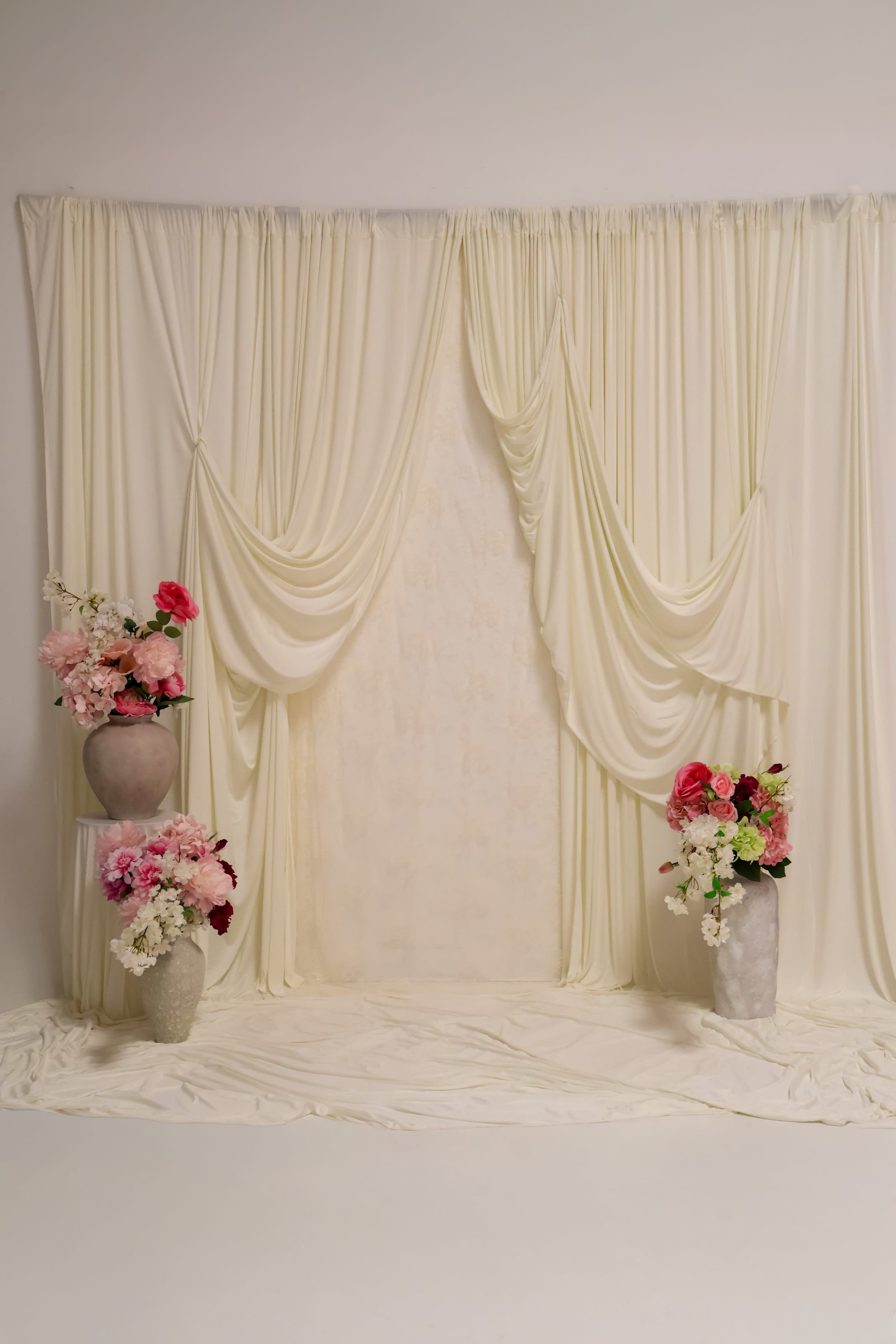 The image shows a staged backdrop with draped white fabric and two floral arrangements in stone vases on either side.