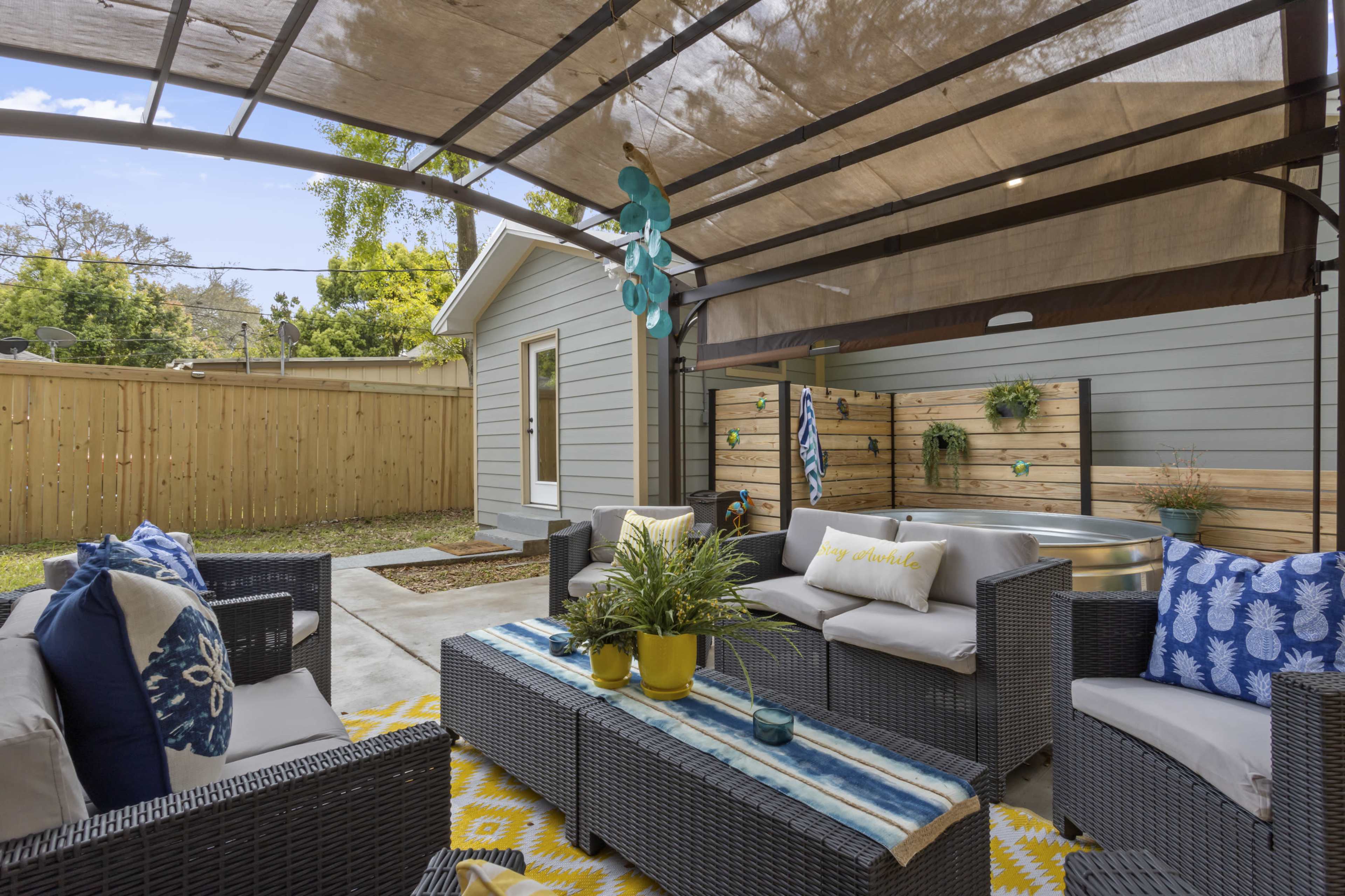 A shaded outdoor living area features a seating arrangement with cushioned chairs, a coffee table, and decorative throw pillows, surrounded by wooden fencing and greenery.