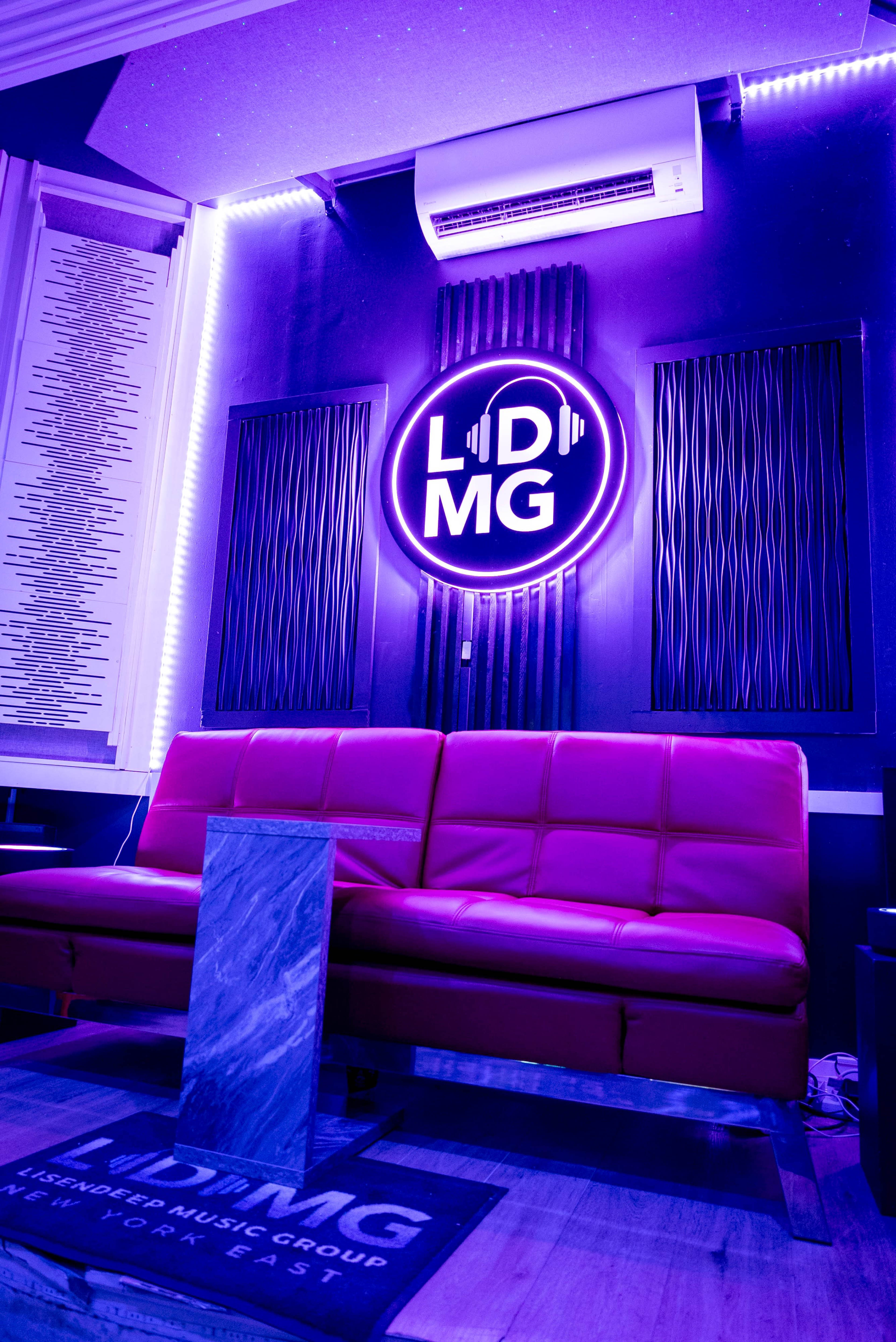 The image shows a modern room featuring a pink couch, a neon sign that reads "LDMG," and acoustic panels on the walls.