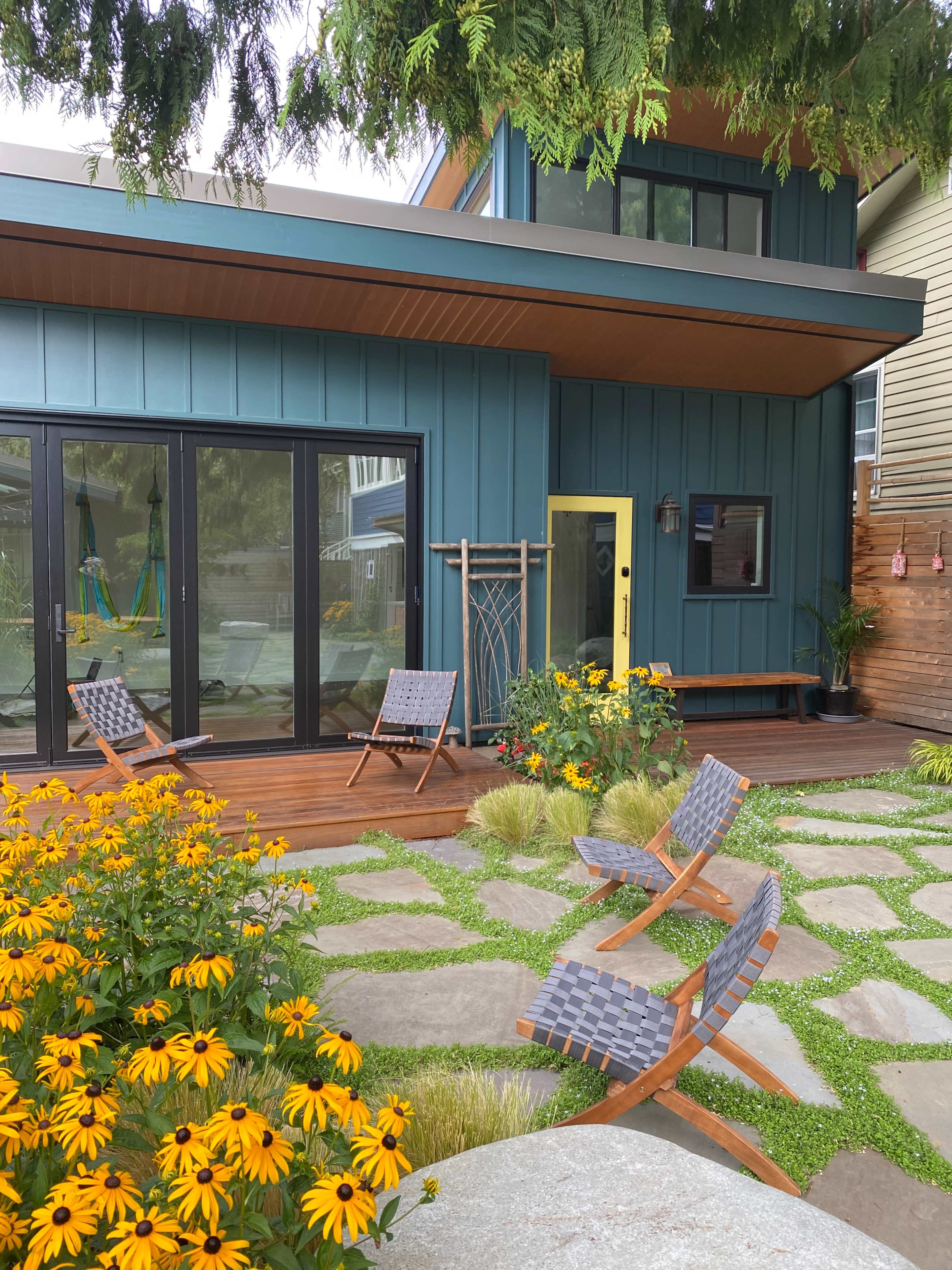 The image shows a modern two-story home with blue siding, large windows, and a yellow door, surrounded by a garden featuring yellow flowers and stone pathways leading to wooden lounge chairs.